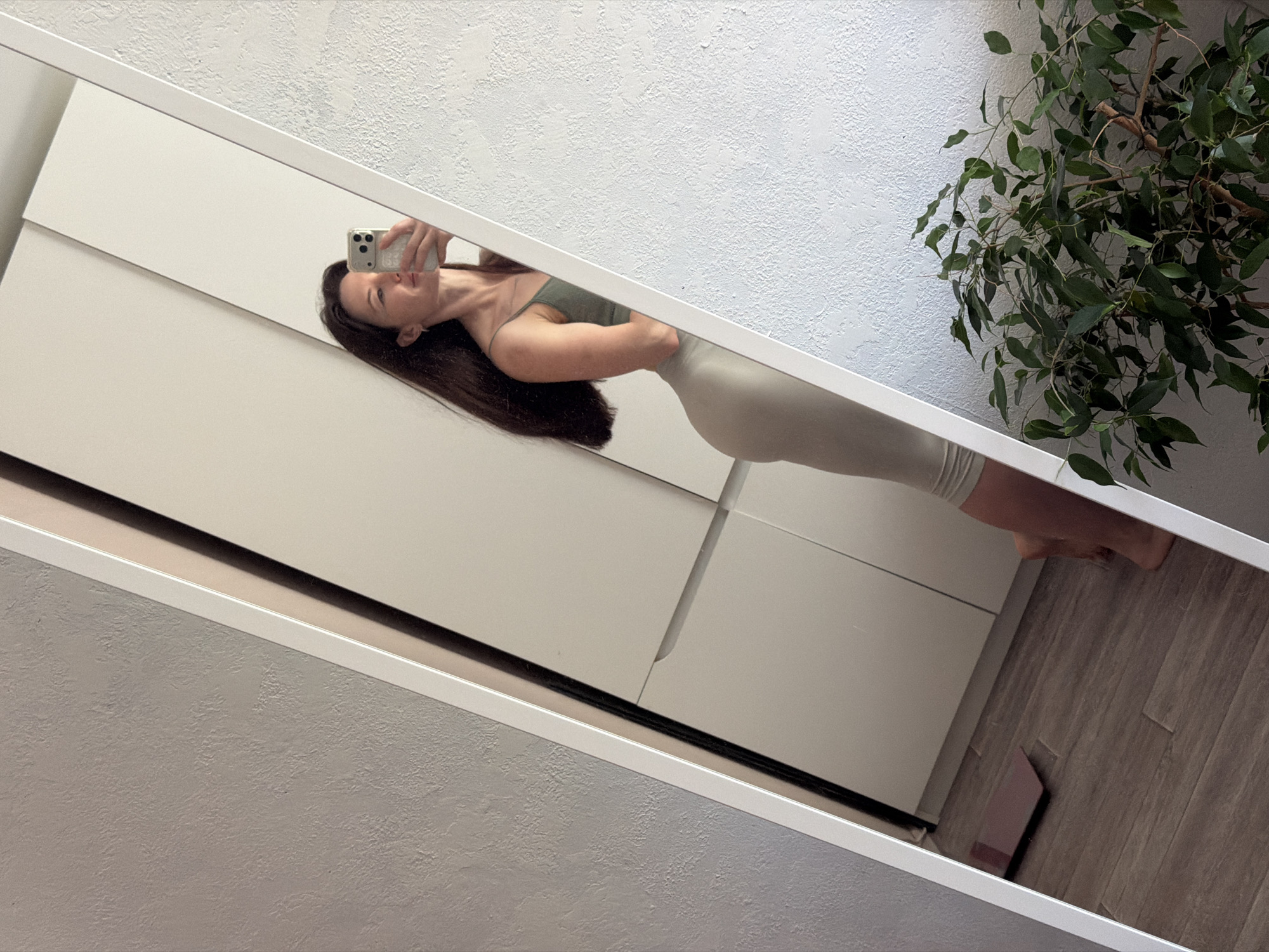 The image shows a woman taking a selfie in a mirror. The woman is wearing a light green tank top and grey shorts. She has long brown hair. She is holding a smartphone in her right hand. The mirror is located in a room with white walls. There is a plant in the upper right corner of the image. The floor is made of wood. It's difficult to determine the exact location of this photo within Perm, Russia without more information.