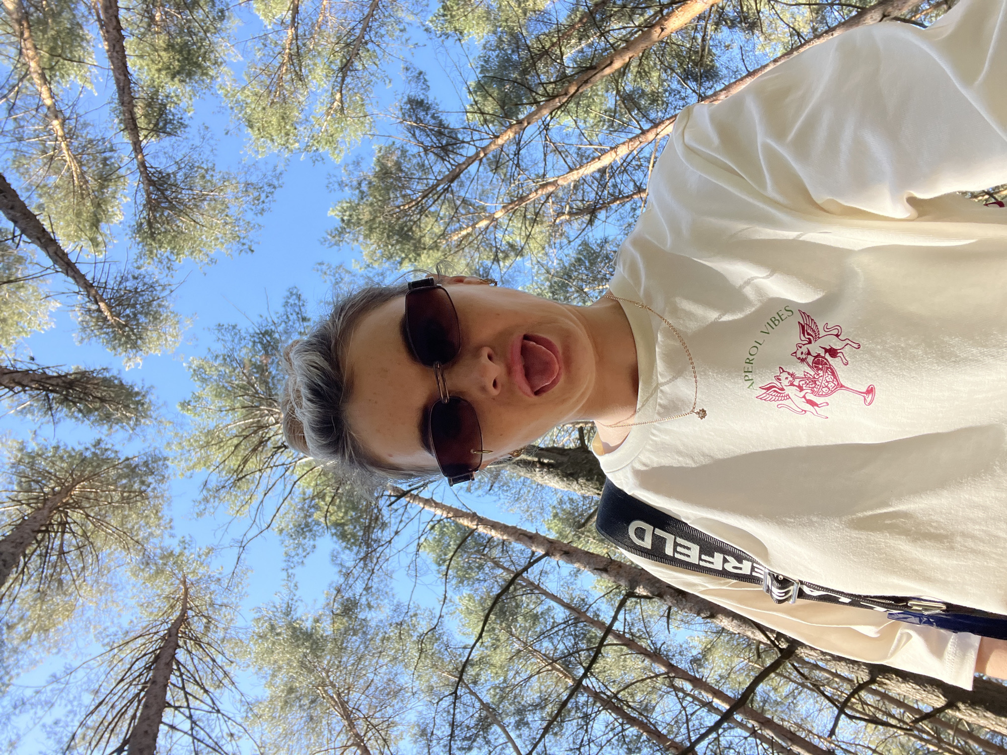 A selfie taken in a forest near Chaikovsky, Russia, angled upwards to include the tops of pine trees and a clear blue sky. The subject is a person with fair skin, short hair styled in a bun, wearing sunglasses and a light-colored t-shirt with a graphic of a cat-like winged creature and a glass, possibly referencing "Aperol Vibes." The person is sticking out their tongue. A black strap with the word "KARLFELD" is visible across the body, indicating a bag or accessory. The environment consists of numerous tall pine trees, their branches extending across the sky, typical of a boreal forest. The lighting suggests a sunny day, with shadows cast by the trees.