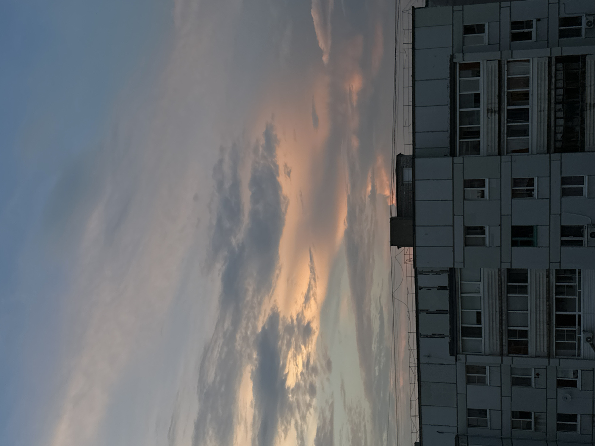 The image captures a serene twilight scene over Chaikovsky, Russia, focusing on the upper facade of a multi-story residential building against a pastel-hued sky. The building, likely constructed from concrete panels, exhibits a grid-like arrangement of windows and balconies. Some balconies appear enclosed, while others are open, with varying states of maintenance visible. Wires and some antennas are present on the roofline, hinting at urban infrastructure. The sky is a gradient of soft blues, grays, and subtle pinks, illuminated by the setting sun, which casts a warm glow on the scattered clouds. No people are visible in the frame, and the overall atmosphere is one of quiet urban stillness at dusk.