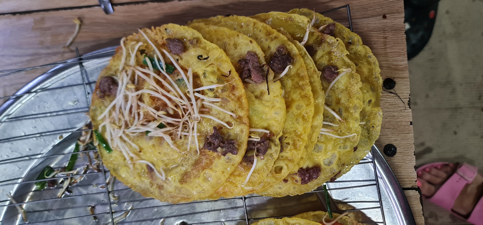 This close-up shot features several freshly prepared Bánh xèo, a traditional Vietnamese savory pancake, likely from a street food stall or a casual eatery in Đà Nẵng, Vietnam.
The main subjects are multiple vibrant yellow, thin, and round pancakes, arranged side-by-side on a silver-colored metal cooling rack with a grid pattern. Each pancake is generously topped with a medley of white bean sprouts, small pieces of dark, reddish-brown meat (likely beef or pork), and finely chopped green onions. The pancakes have a slightly uneven, crispy-looking texture, indicative of being freshly fried.
Beneath the cooling rack, a circular metal tray or plate is visible, suggesting that the pancakes are either draining excess oil or waiting to be served. The entire setup rests on a light brown wooden surface, possibly a table or counter. In the blurred background, to the right, a person's foot wearing a bright pink slide sandal is partially visible, reinforcing the informal and possibly outdoor setting, such as a local market or a bustling food street.
The image captures the moment the food is ready for consumption, emphasizing its fresh ingredients and traditional preparation. No visible text is present in the image.