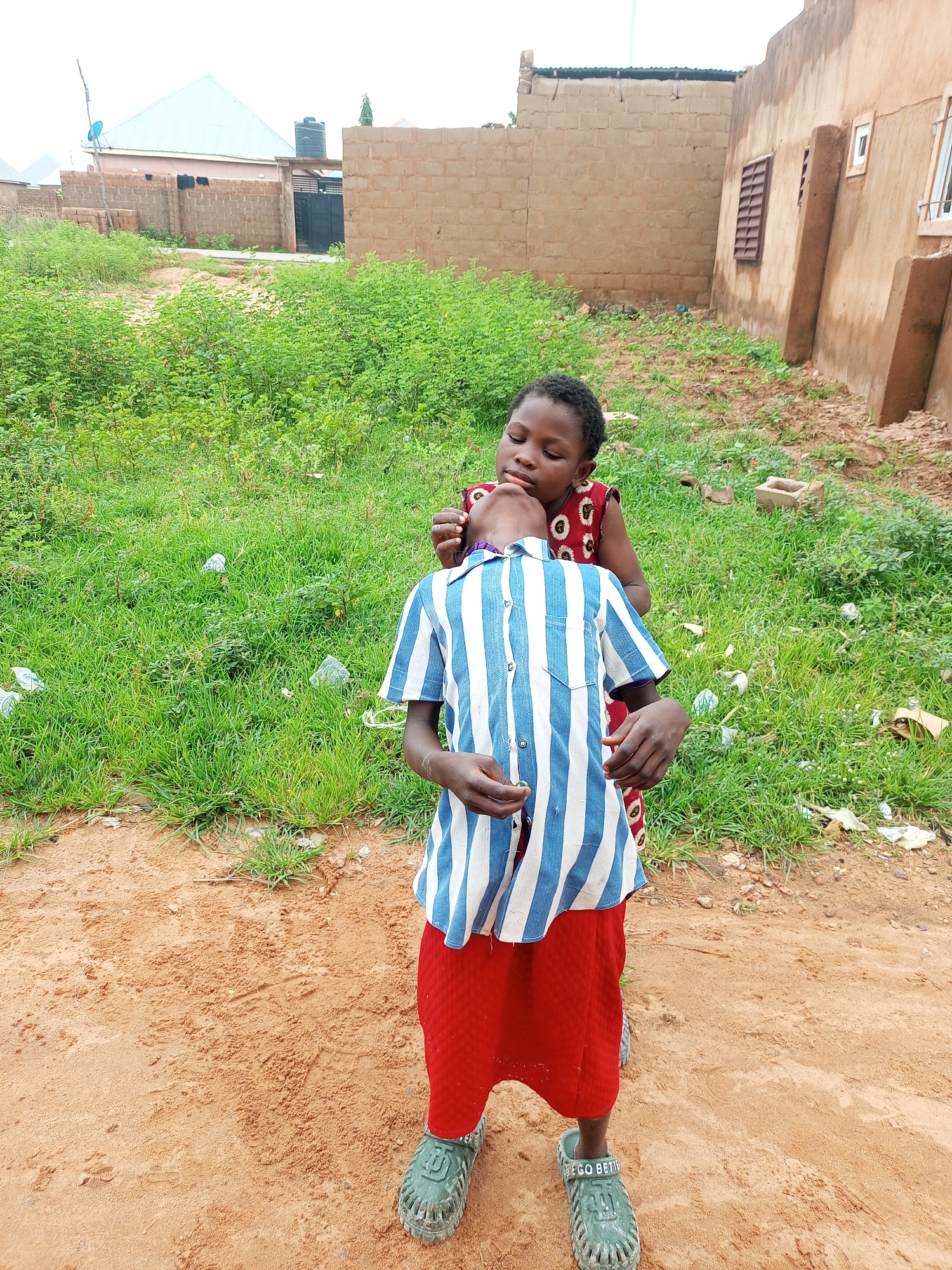 Two young children are standing in a dirt lot in front of a brown, unfinished building. The child in the foreground is wearing a blue and white striped shirt and a red skirt, with green sandals. The child behind them, wearing a red and white patterned dress, is leaning forward and touching the first child's chin. The background features overgrown green foliage and more buildings. The scene appears to be in a rural or developing area.