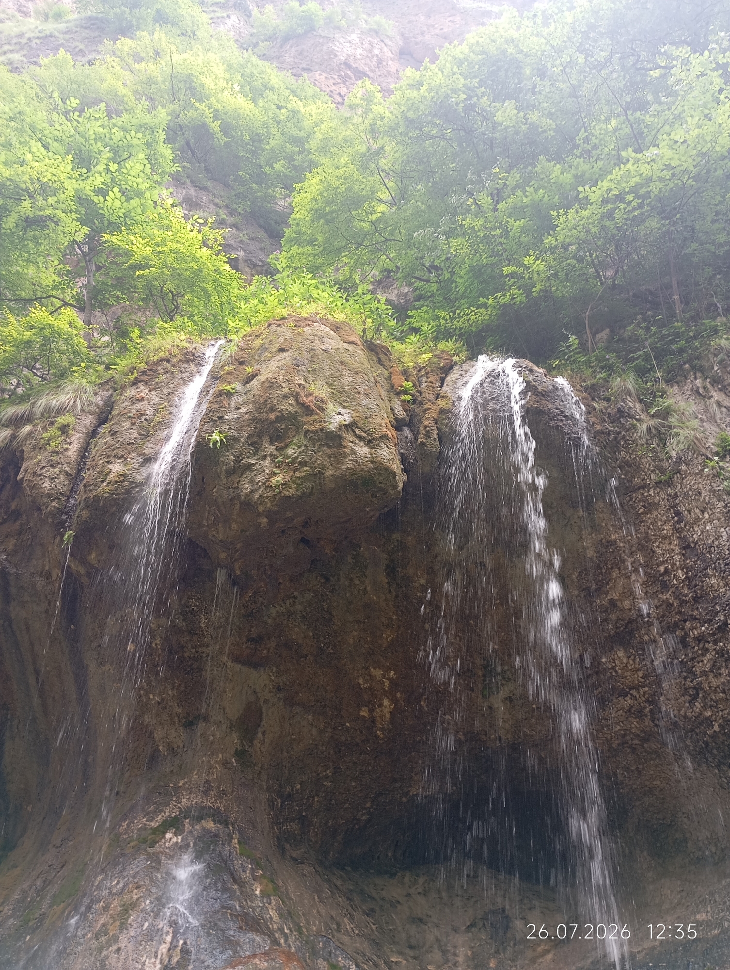 This is a low-angle, vertical, outdoor shot of a rocky cliff face with a waterfall cascading down it. Lush green trees and foliage are visible at the top of the cliff, suggesting a natural, possibly mountainous environment. The water from the waterfall is creating streaks of white against the dark, textured rock. The lighting appears to be overcast or diffused, casting a soft glow over the scene. The image is dated 26.07.2026 12:35, indicating a specific point in time. The location is identified as Khushto-Syrt, Russia. No people or animals are visible in the frame.