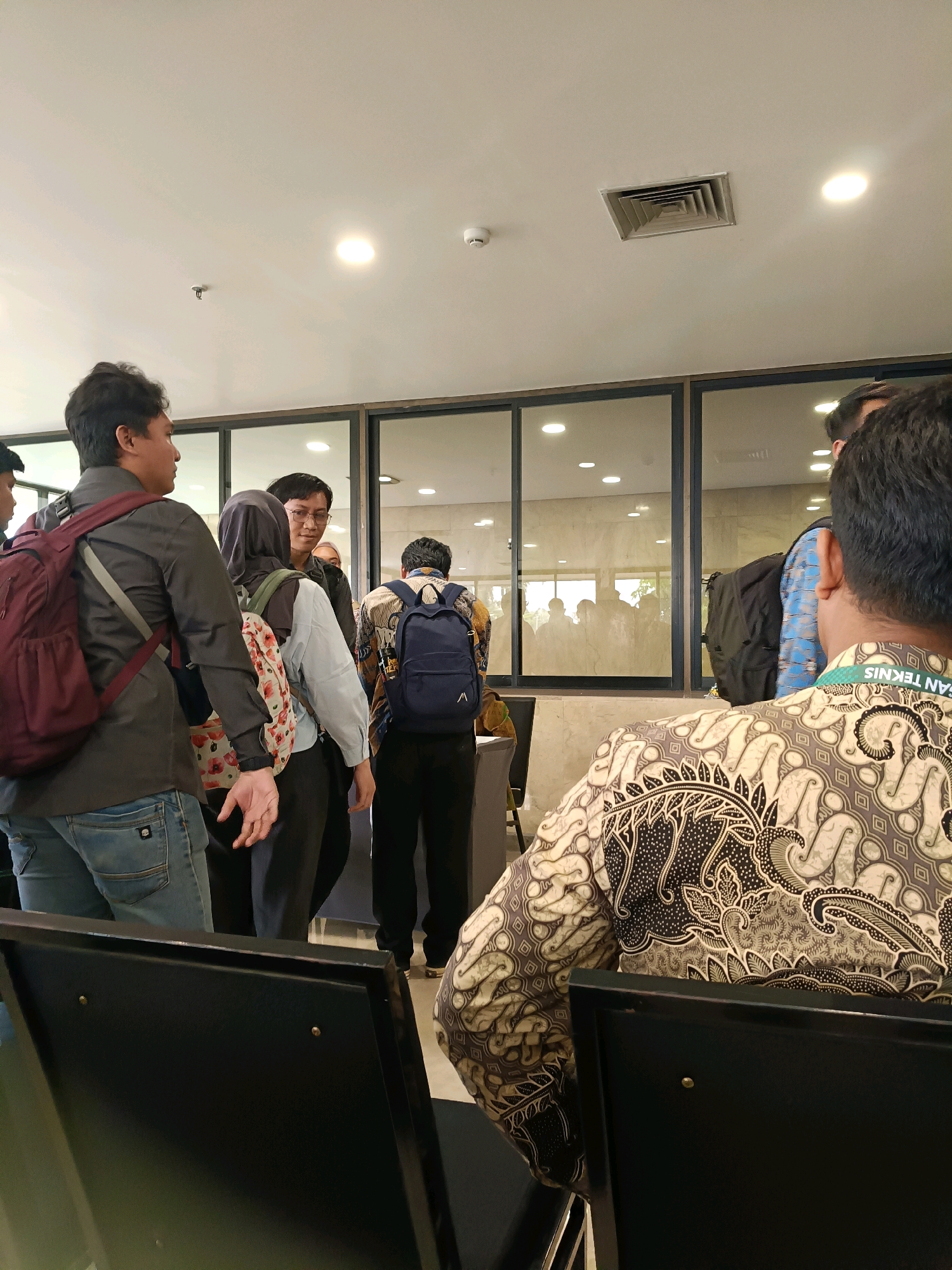 A group of people, mostly young adults, are gathered in an indoor space that appears to be a waiting area or hallway. The setting has modern elements like glass partitions and recessed ceiling lights. Several individuals are wearing backpacks, suggesting they might be students or attending an event. One person in the foreground is wearing a batik patterned shirt, a traditional Indonesian garment. Another person is wearing a lanyard with a badge, indicating they might be an employee or participant in an organized activity. The environment suggests a public or semi-public indoor space. The lighting is bright, typical of an office or institutional building. The overall impression is of a casual gathering or a moment of waiting. Based on the presence of batik, modern architecture, and the general demographic, it is plausible this scene is located in North Jakarta, Indonesia, possibly within a university or government building.