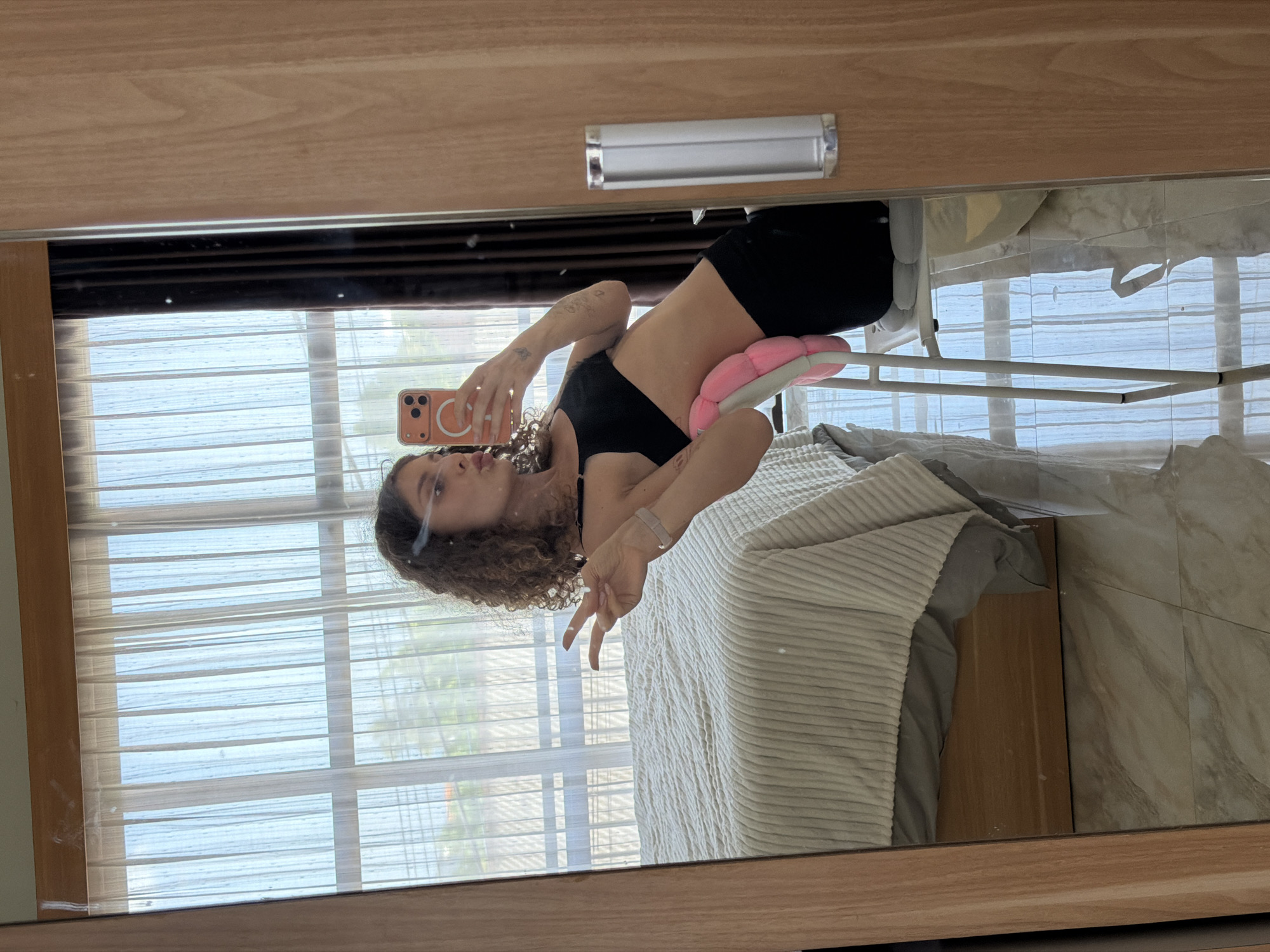 A woman with curly brown hair is taking a mirror selfie in a room. She is wearing a black sports bra and black leggings. Her left hand is raised, making a peace sign. She is sitting on a white stool with a pink cushion behind her lower back. In the reflection, a bed with a white comforter is visible, along with a window with sheer white curtains. The floor is tiled with a light marble pattern. The setting appears to be a bedroom or a similar residential space, likely located in North Kuta, Indonesia, given the style of the window and furniture.