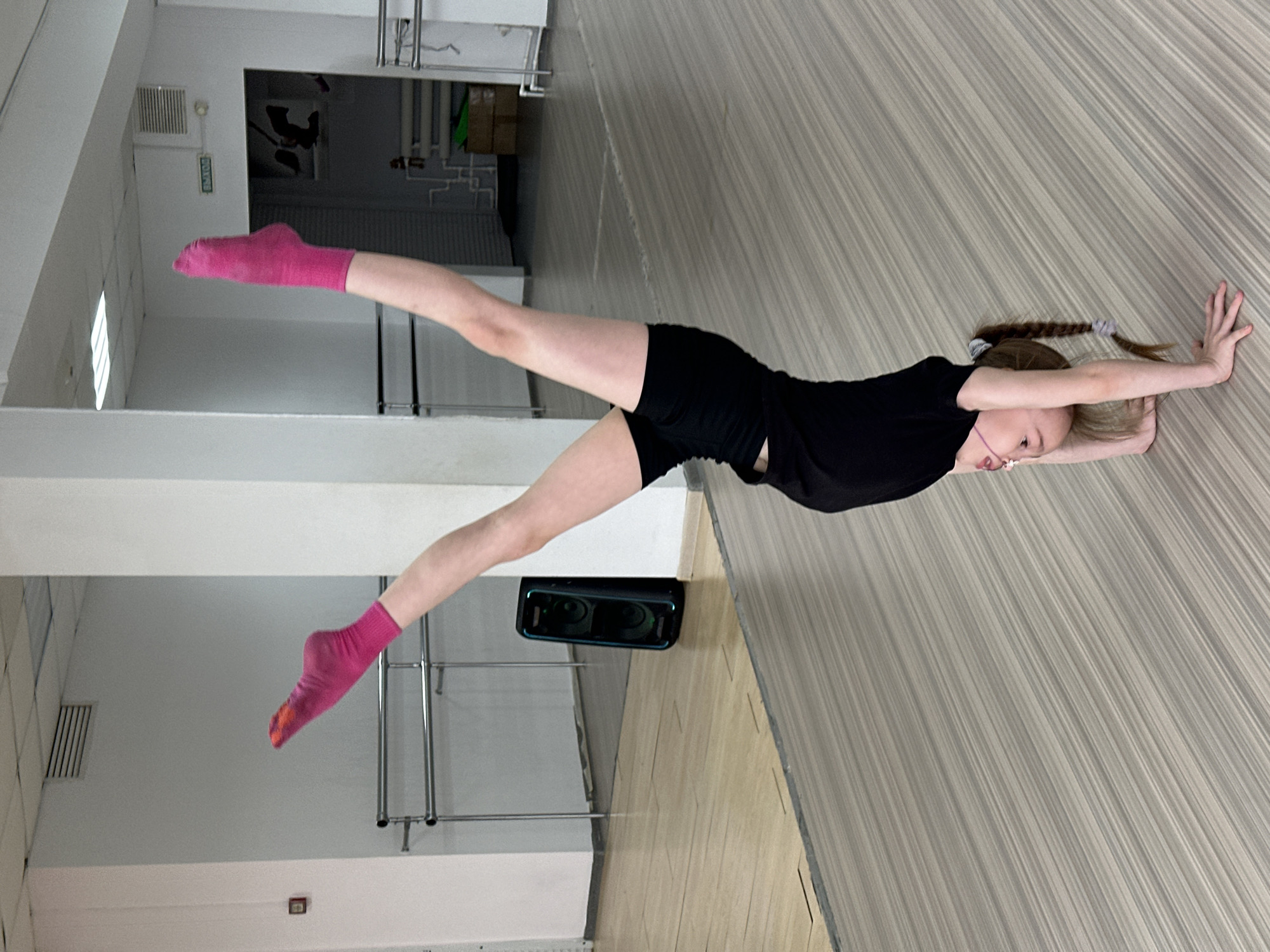 A young female is captured mid-stretch in what appears to be a dance or exercise studio. She is dressed in black shorts and a black top, with pink socks on her feet. Her body is horizontal, parallel to the floor, with one leg extended upwards and the other extended downwards. Her arms are stretched out in front of her. The background includes a wall with a light-colored striped pattern, presumably the studio floor. The room has elements like white walls, metal bars, and a speaker. There is no verifiable evidence indicating that the photograph was taken in Votkinsk, Russia.