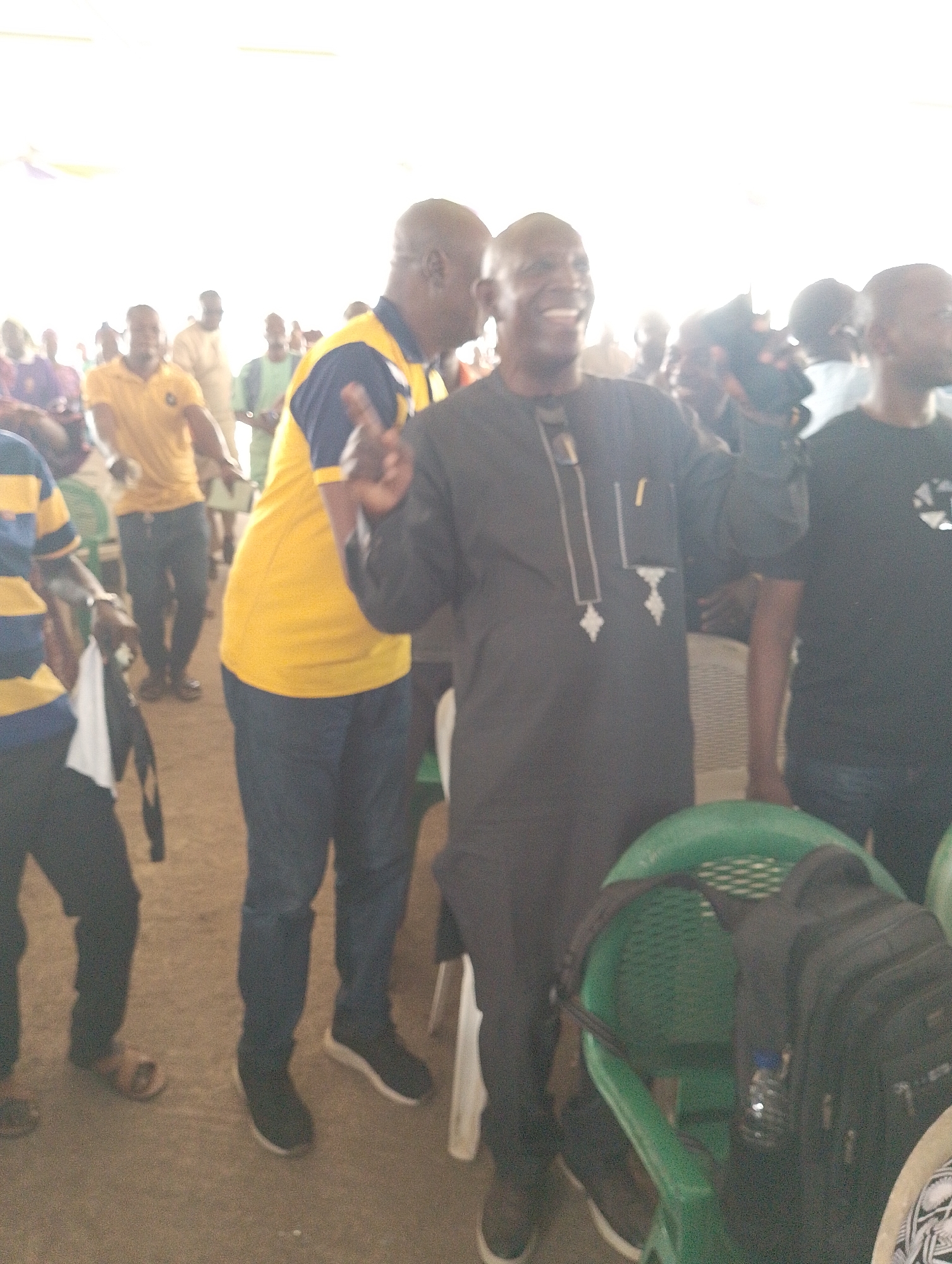 This image captures a bright, somewhat overexposed scene of a social gathering, likely indoors or under a canopy. The main subject is a dark-skinned man with a bald head, positioned centrally and smiling broadly, revealing his teeth. He is wearing a dark grey/black long-sleeved tunic, possibly an agbada, featuring distinct white embroidery down the front and on the cuffs. He holds up his right hand with two fingers extended, forming a 'peace' or 'victory' sign. A small, light-colored rectangular patch is visible on his upper left chest, possibly a pin or ID, but no text is legible.
Behind him and slightly to his left, another man with a bald head is visible, wearing a yellow polo shirt with navy blue sleeves and dark trousers. He is partially obscured and facing away from the camera. On the far left, a person wearing a striped blue and yellow top is partially visible, with a white shirt draped over their arm. To the right of the central figure, another individual in a black t-shirt is visible, though their face is cropped out.
The setting appears informal, with a light-colored ground, possibly packed dirt or concrete. Green plastic chairs are scattered around, one of which in the foreground holds a black backpack and a small water bottle. The lighting is very bright, suggesting either strong natural light or powerful artificial illumination. The mood conveyed by the central smiling figure is one of happiness and enthusiasm. No discernible text or specific location cues are visible.