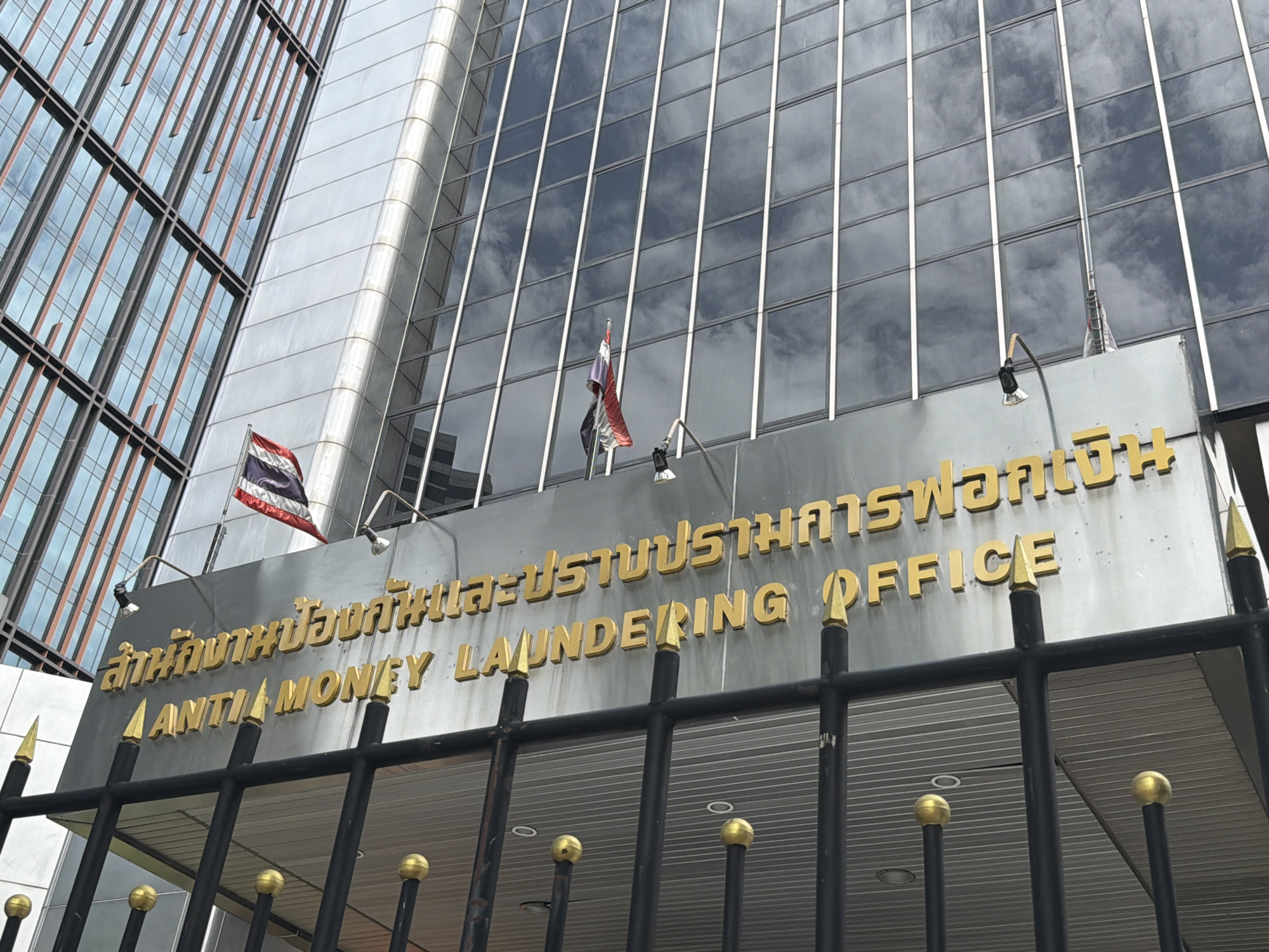 The image shows the exterior of a modern building with a glass facade in Bangkok, Thailand. The building is identified by prominent golden lettering that reads "ANTI MONEY LAUNDERING OFFICE" in English and what appears to be Thai script above it. Two flags are visible on the building's exterior: a Thai flag and another flag that is partially obscured. The building is bordered by a black metal fence with gold finials in the foreground. The overall scene depicts the official headquarters of the Anti-Money Laundering Office in Bangkok.