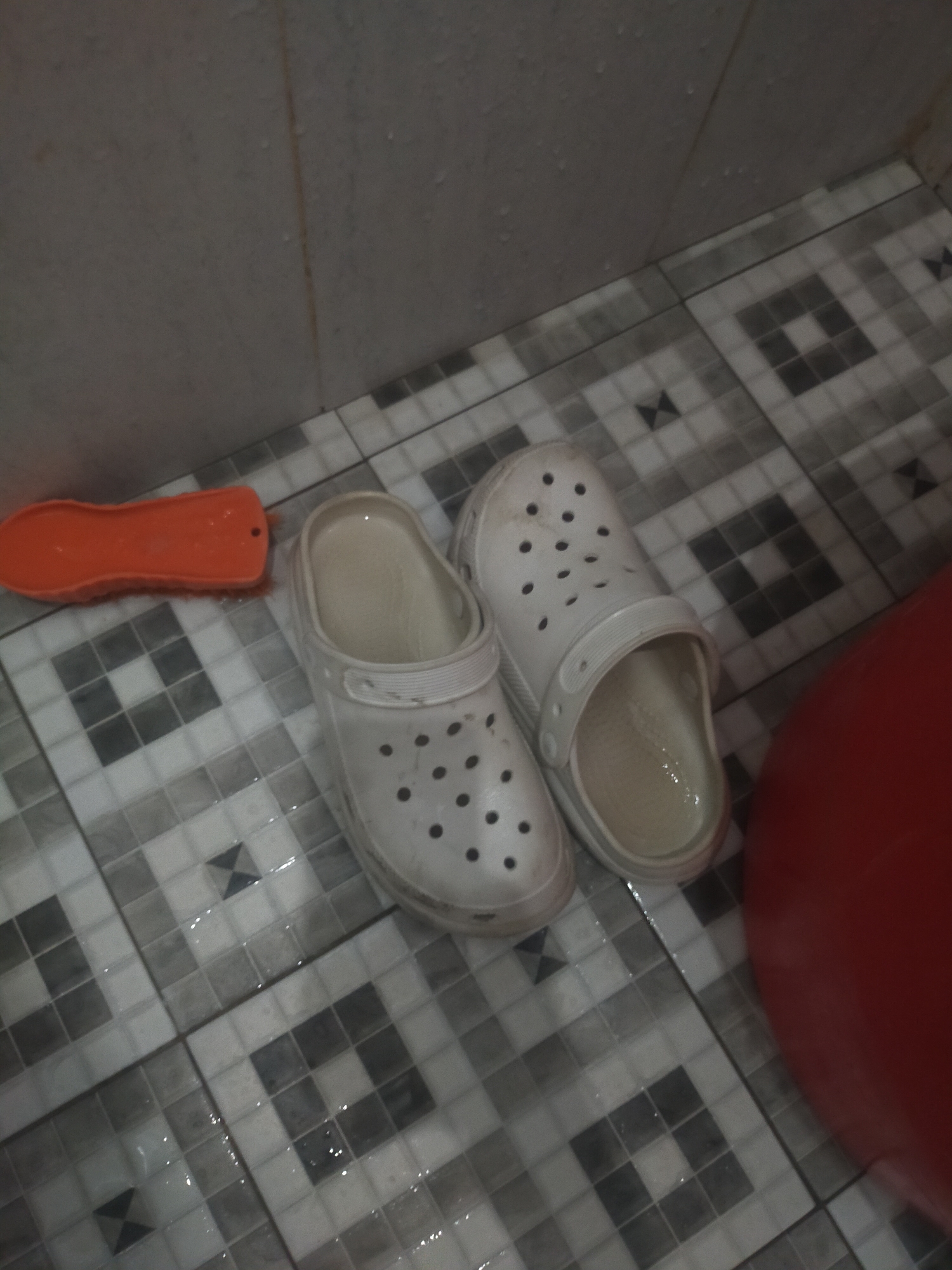 In a bathroom with tiled flooring, two white Crocs are positioned side-by-side. An orange scrub brush lies to the left of the shoes. A large, red cylindrical object, possibly a bucket, is visible to the right of the Crocs. The floor appears wet. The scene is indoors, consistent with a residential bathroom in Tegalandong, Indonesia.