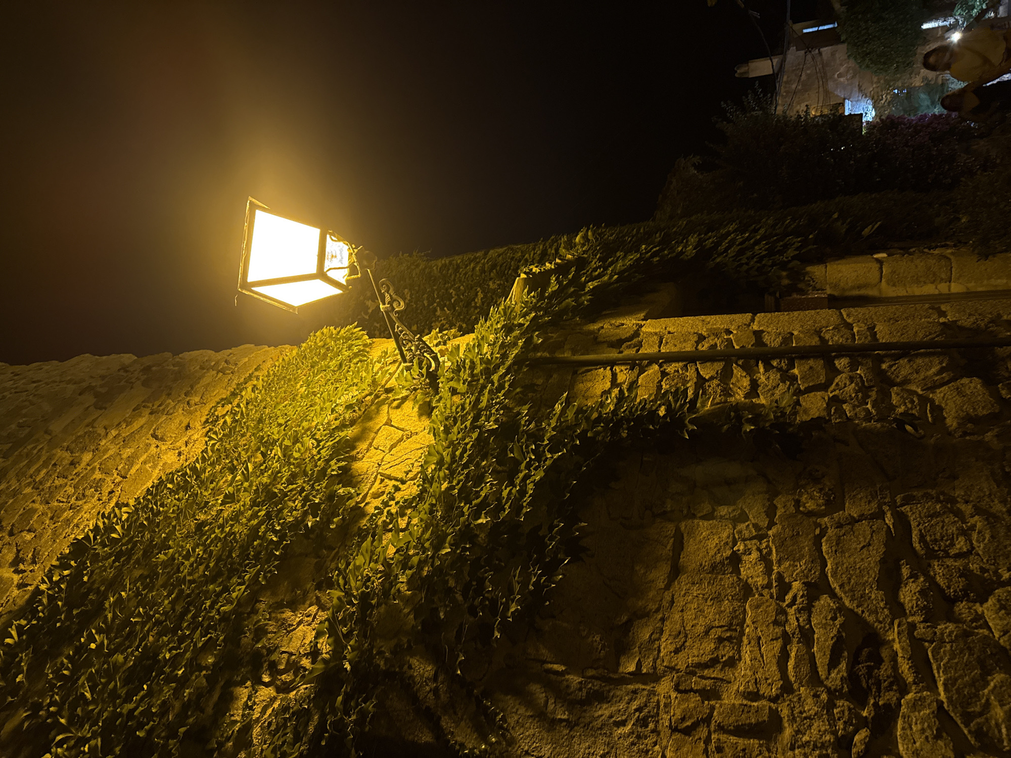 A vintage-style streetlamp illuminates a stone wall covered in ivy at night in Tossa de Mar, Spain. The lamp, with a white rectangular shade, casts a warm yellow glow on the textured stone and green foliage. The wall itself is constructed from large, rough-hewn stones, suggesting an old building. The darkness of the night sky provides a stark contrast to the illuminated area. In the background, a narrow street is visible, with hints of other buildings and vegetation, and two individuals walking away from the camera.