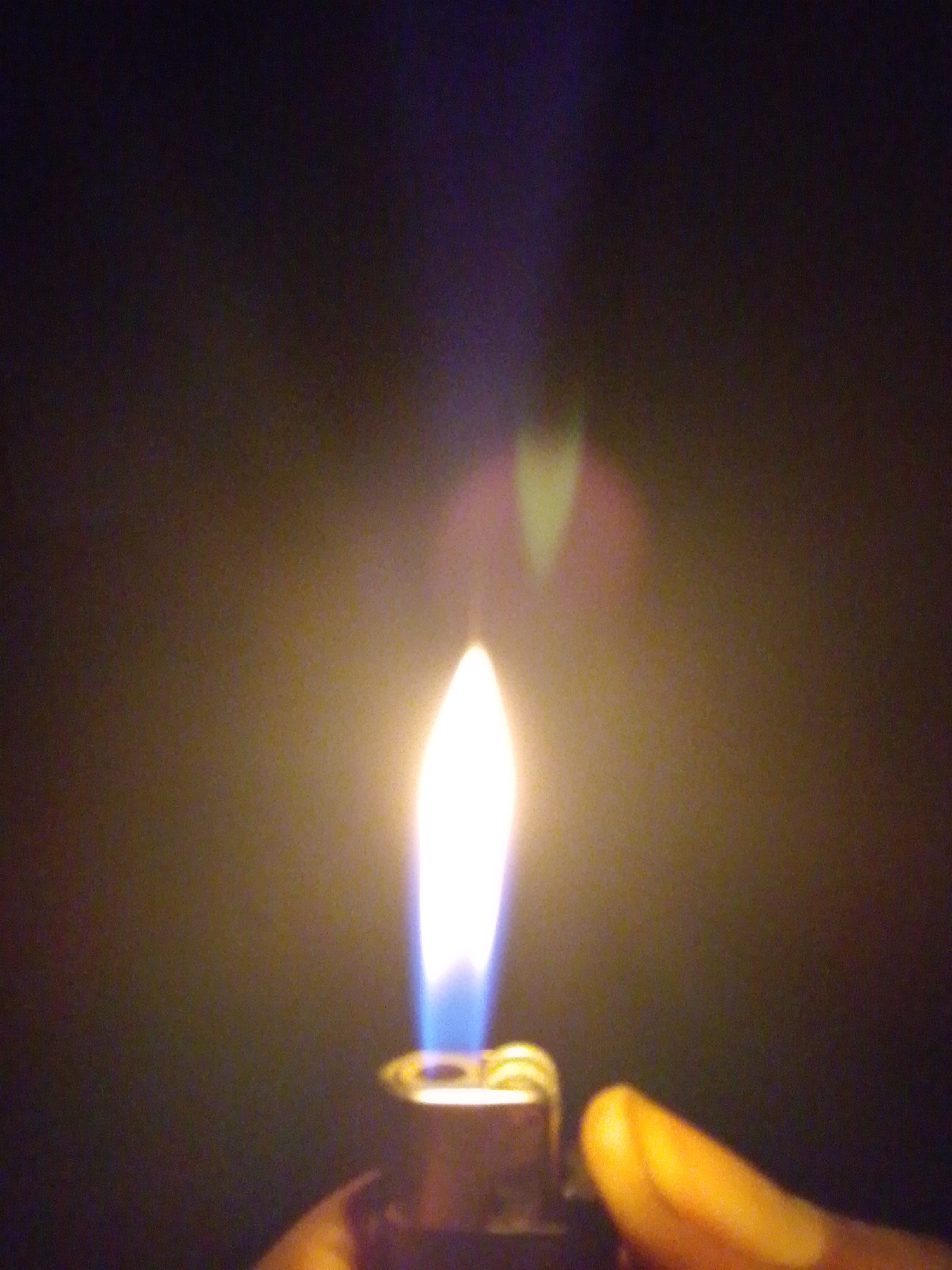 The image is a close-up of a lighter with a flame, held in a person's hand. The background is dark and out of focus. The flame is bright white at the tip and transitions to blue at the base, indicating a gas flame. The lighter appears to be made of metal and is partially visible at the bottom of the frame. A person's thumb is visible on the right side, holding the lighter. The scene does not provide any information about the city of Serti, Nigeria, or its specific location within it.