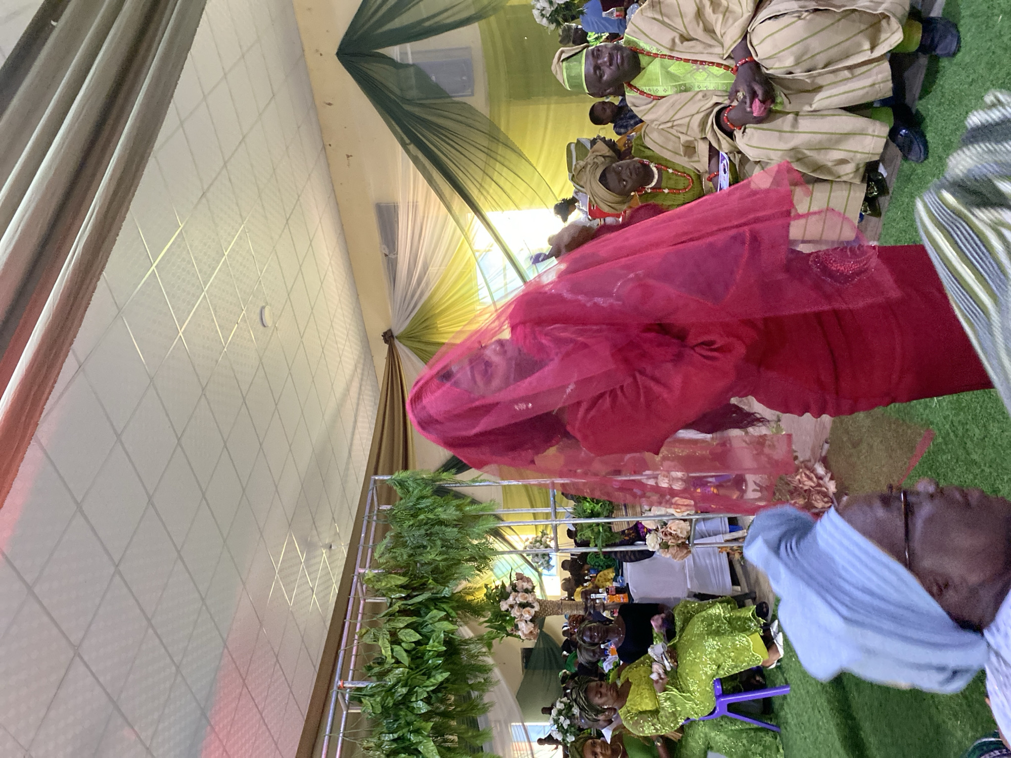 The image shows an indoor event in Kuru Karama, Nigeria, with a mix of decorative fabrics in shades of green, yellow, and white draped from the ceiling. A floral arrangement hangs from a silver metal frame adorned with foliage. Seated guests, some wearing traditional Nigerian clothing, are visible. A woman in a red dress with a red veil is a primary subject. A red carpet is partially visible.