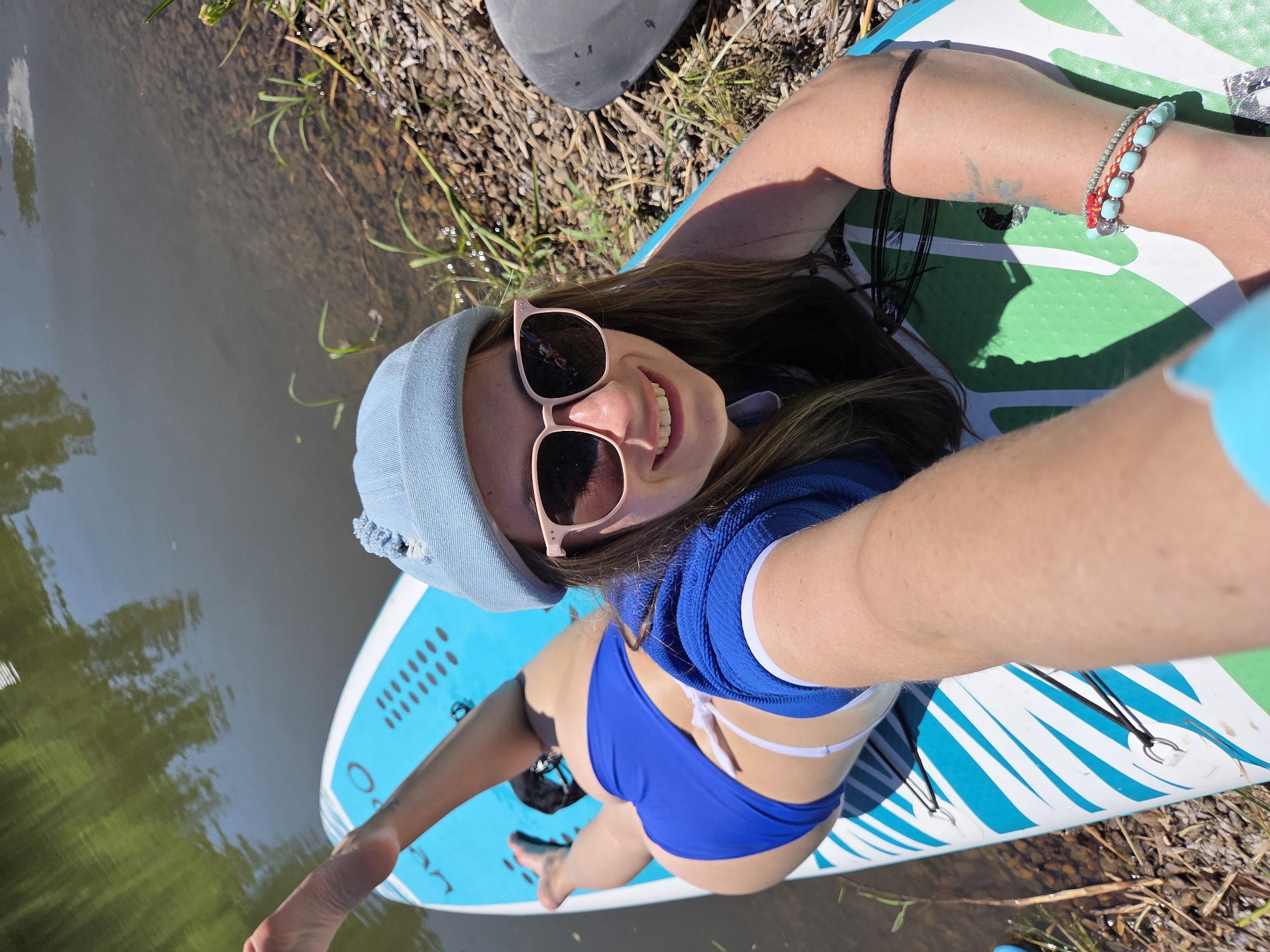 A woman is lying on a paddleboard in a body of water. She is wearing a blue beanie, pink sunglasses, and a blue bikini. Her left leg is raised in the air. The paddleboard is blue and white with green accents. The water is dark and reflects the trees on the shore. The shore is covered in gravel and dry grass. The image is taken from a high angle, looking down at the woman. It is a sunny day and the woman is smiling. The specific location in Birobidzhan, Russia is not discernible from the image.