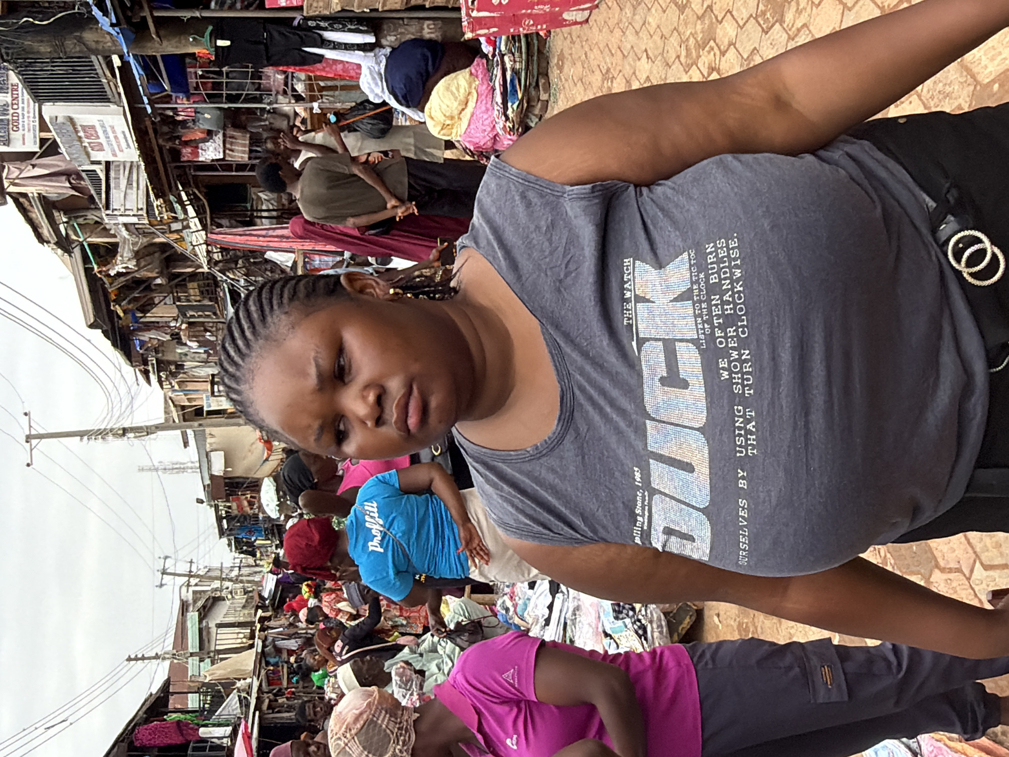 The image shows a woman wearing a gray tank top with the word "BUCK" printed on it. She is standing in an outdoor market, which appears crowded and bustling with people and merchants displaying their wares, predominantly clothing. Structures visible in the background suggest an urban environment with stalls or shops lining the street. There are utility poles in the distance and a building with a sign that reads "GOLD CENTRE". A man is wearing a blue shirt printed with "Proffill." Another person is wearing a pink shirt. The market appears to be located in Jos, Nigeria.