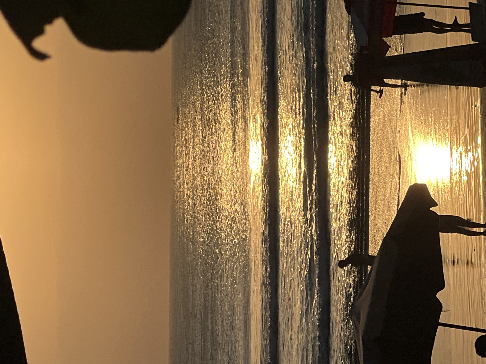 The image depicts a beach scene at sunset in Kuta, Indonesia. Silhouettes of people and beach umbrellas are visible on the sand. The sun is setting over the ocean, casting a warm, golden glow on the water and the sky. The waves are gentle, and the overall atmosphere is tranquil. A small boat is visible in the distance on the water.