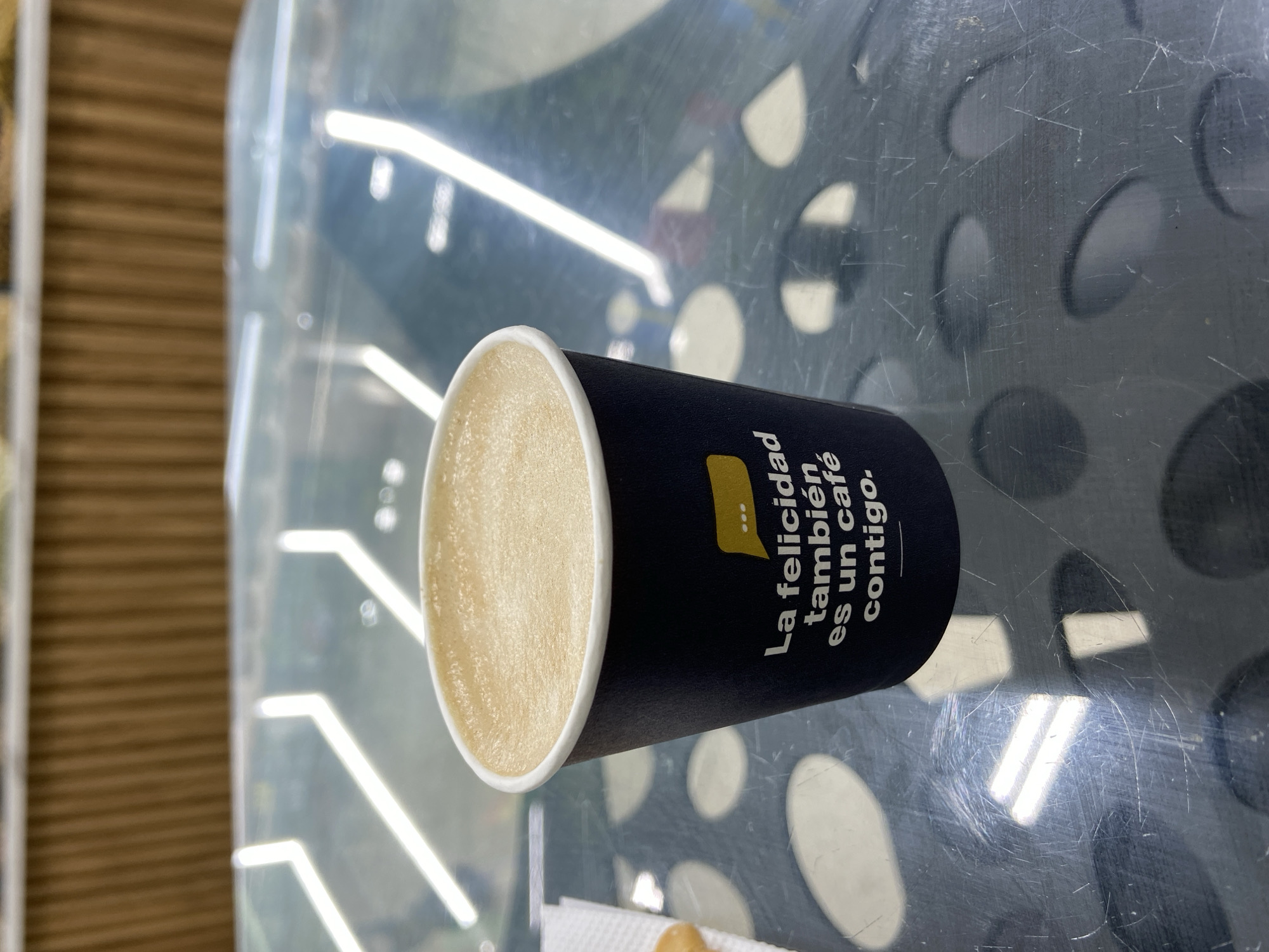 A dark blue paper cup filled with a light-colored coffee drink topped with foam sits on a reflective, speckled metallic surface. The cup has white text that reads "La felicidad también es un café contigo." The background is blurred, showing a light brown ribbed surface and a bright, abstract reflective pattern on the surface the cup is resting on. The image is taken from a medium-close, slightly overhead angle.