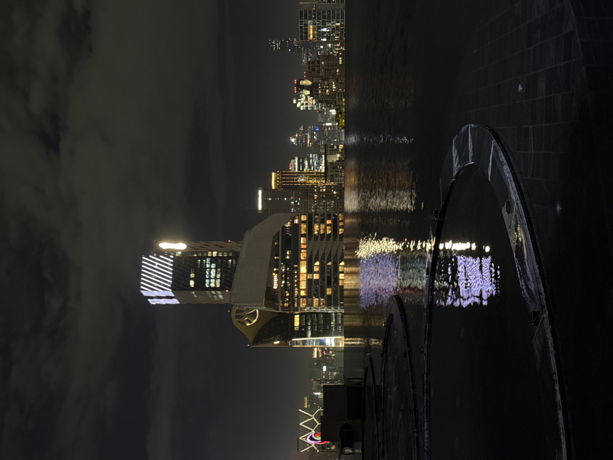 This is a nighttime photograph of a cityscape with a focus on modern architecture and water reflections. The primary subject is a cluster of illuminated skyscrapers, suggesting an urban environment. One prominent building in the center features a distinctive gold emblem and a white, patterned lighting design on its upper section. Other buildings in the background display various lighting patterns and colors, indicative of a bustling city. In the foreground, a dark, reflective water surface, possibly a pool or a canal, mirrors the city lights, creating a dual image. The water is bordered by dark, tiled edges, hinting at an elevated viewpoint such as a rooftop. The sky is dark with some cloud cover. The overall impression is one of a vibrant, modern city at night, with the interplay of light and water creating a dramatic visual effect. While the image itself doesn't explicitly state the location, the architectural style and the presence of illuminated skyscrapers are consistent with major metropolitan areas. For Bangkok, Thailand, this scene is highly characteristic of the Sukhumvit area, known for its modern high-rises, shopping centers, and entertainment venues, particularly around the Asok intersection where buildings like the Grand Hyatt Erawan Bangkok and Terminal 21 are located, known for their distinctive architecture and illumination.