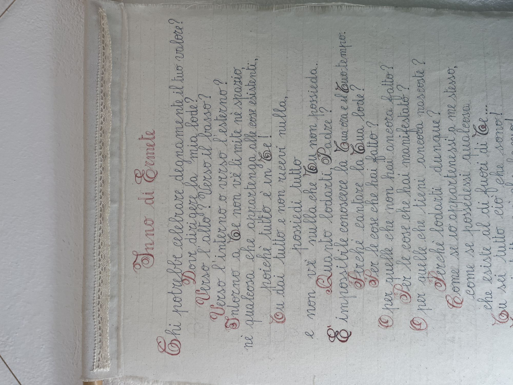 The image shows a handwritten poem titled "Inno di Ermete" (Hymn of Hermes) on what appears to be a piece of fabric, possibly linen, trimmed with lace. The text is written in cursive script with some decorative flourishes, using two colors - a dark color for the majority of the text and a lighter reddish color for emphasized words and the initial letters of each stanza. The poem is written in Italian.