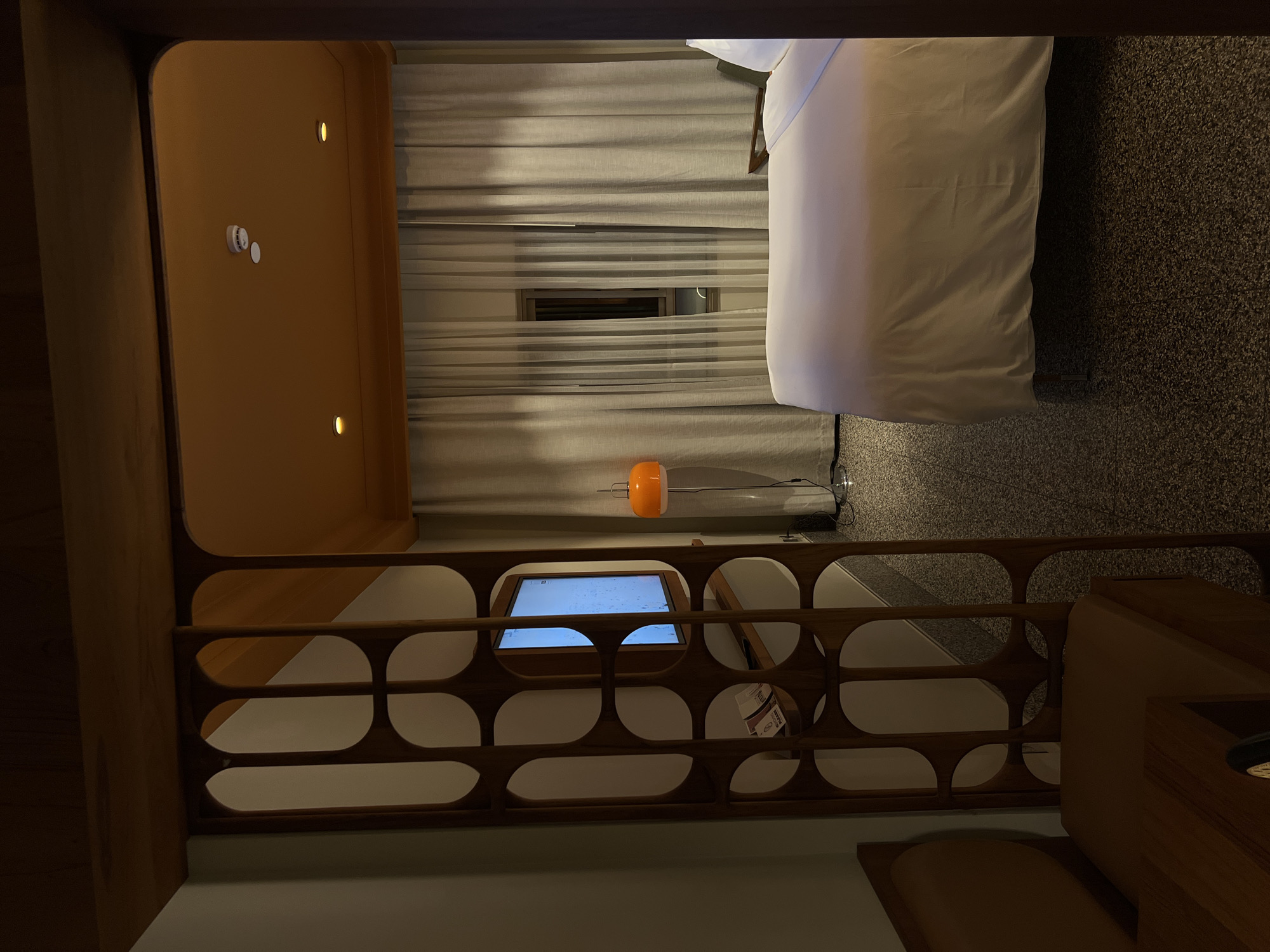 This image captures a multi-level, compact living space, possibly a modern hotel room or capsule accommodation, characterized by its warm wooden accents and contemporary design. The scene appears to be set at night or in a darkened environment, illuminated by interior lighting.
On the upper level, a bed with clean white bedding is neatly made on the right side of the frame. To the left of the bed, a window is mostly covered by light-colored horizontal blinds, with a small section open in the middle, revealing only darkness beyond, reinforcing the nighttime ambiance. An orange, spherical table lamp with a slender metallic stand sits on the dark, speckled carpeted floor near the window.
The lower level is separated from the upper by a distinctive wooden railing featuring a repeating pattern of rounded, rectangular cutouts, suggesting a mid-century modern aesthetic. Behind this railing, a screen or monitor displays a glowing light blue interface with some faint white patterns, possibly a control panel for the room or an information display. On a wooden wall to the far left of the upper level, two small, circular recessed lights provide illumination. A white smoke detector and a smaller white disc (likely another sensor) are also mounted on this wooden wall. Another section of wooden furniture or wall is visible in the bottom right corner of the frame.
There are no people present in the image. The artificial lighting, primarily from the recessed lights and the orange lamp, creates a cozy and intimate atmosphere within the efficiently designed space. No specific legible text is visible in the image.