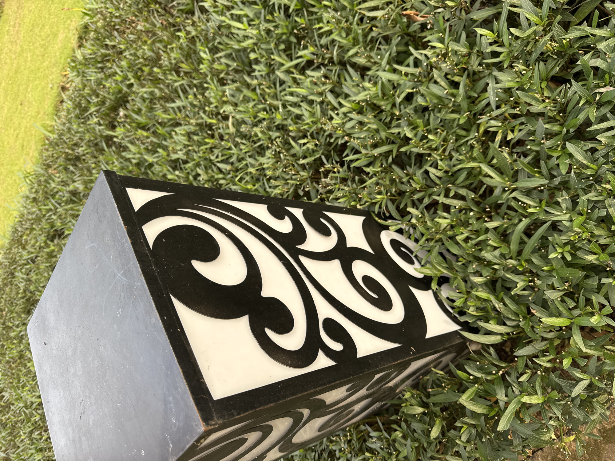 A gray and black cuboid structure with white panels featuring black scrollwork stands on a patch of green foliage. The foliage appears to be a type of small-leaved shrub. The scene takes place outdoors, likely in Kuta Selatan, Indonesia. The cuboid structure may be a lamp or decorative object. There are no people visible in the image.