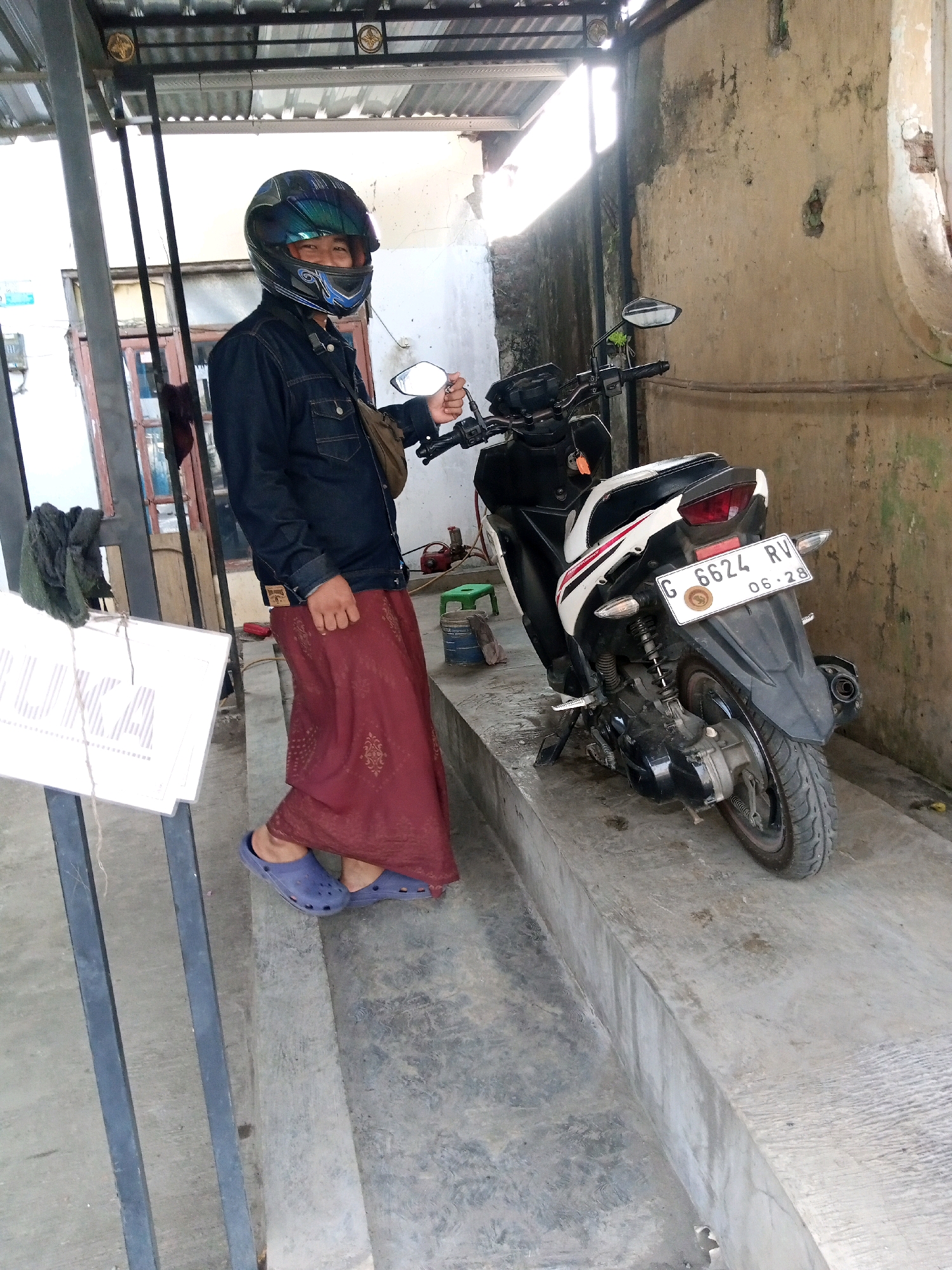 A man wearing a dark denim jacket, a maroon sarong, and blue clogs stands next to a white and black motorcycle. He also wears a blue and green motorcycle helmet with a reflective visor. The man is holding the motorcycle's handlebar with his right hand. He has a light brown bag slung over his left shoulder. The motorcycle has a white license plate with black lettering that reads "G 6624 RV" and "06-28". The scene appears to be an outdoor area with a concrete floor and a wall behind the motorcycle. There is a metal structure with a corrugated roof above the man. A white sign with the word "LUXCA" is visible to the left of the man, attached to a black metal pole. The location is identified as Proyonanggan Tengah, Indonesia.
