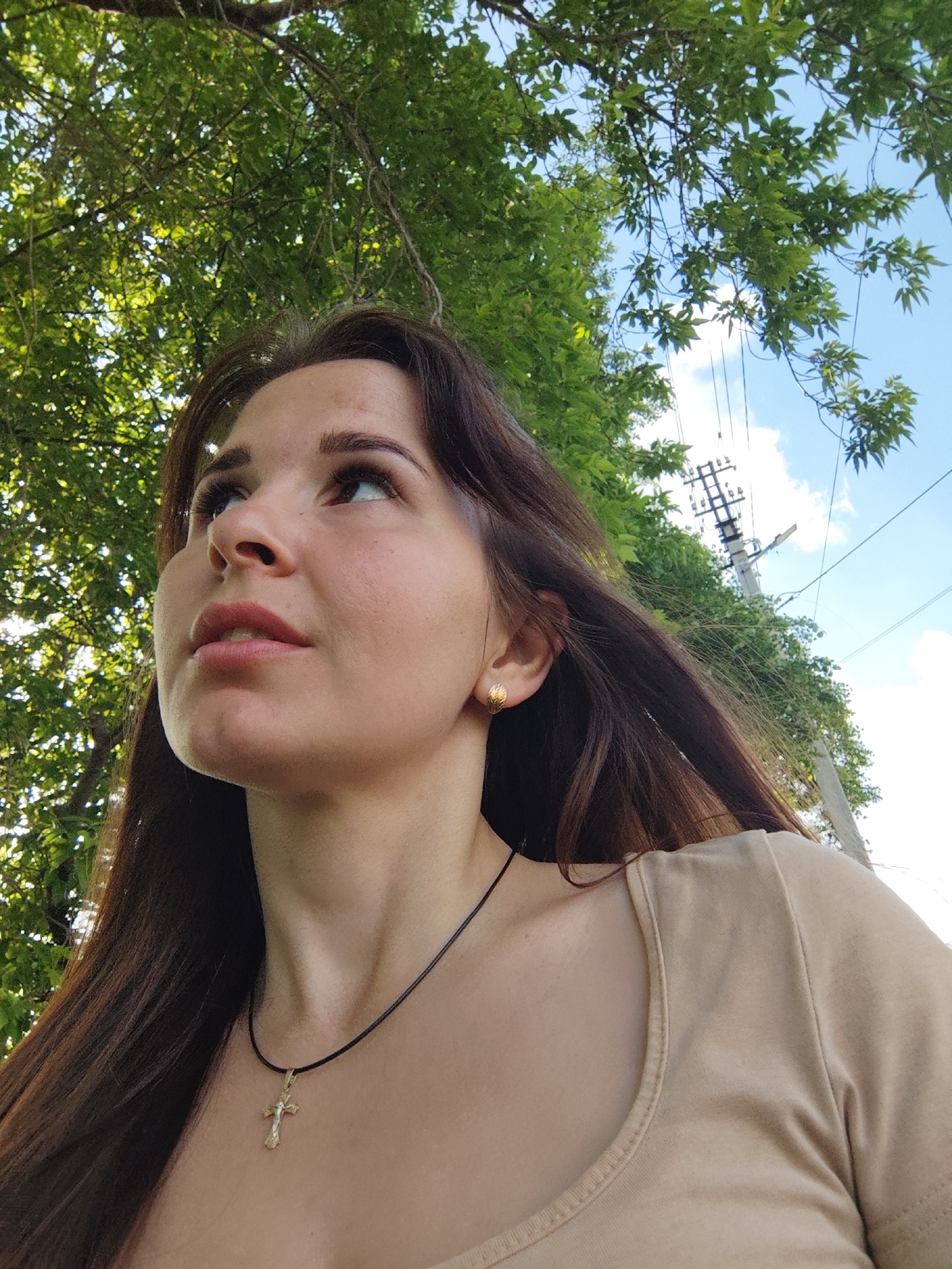 A low-angle outdoor portrait features a young woman with long, dark brown hair, looking upwards towards the sky. Her face is seen in profile, with a contemplative expression. She wears a light beige top with a scooped neckline and a black cord necklace adorned with a small, gold-toned cross pendant. A small, gold stud earring is visible in her right ear.
The background is filled with lush green foliage of trees, through which bright sunlight filters, creating a dappled effect. Above the trees, a clear blue sky with scattered white clouds is visible. To the right, a utility pole with power lines and insulators can be seen against the sky. The image captures a bright, sunny day. The setting appears to be an outdoor natural environment, possibly a park or a roadside area, consistent with the location context of Фоки, Russia. There is no visible text in the image.