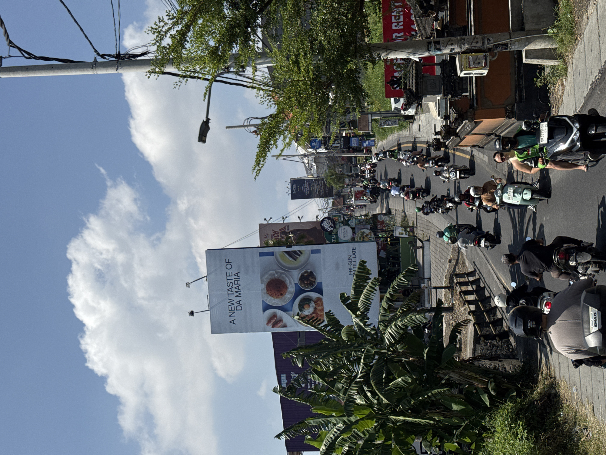 The image captures a bustling street scene in North Kuta, Indonesia, under a clear, bright blue sky with white clouds. The perspective is slightly elevated, looking down a busy road filled with a long queue of motorbikes.
Numerous people are visible, mostly riding scooters, wearing helmets and casual clothing. One rider in the mid-ground is distinctly seen wearing a green vest. The street is dominated by a variety of motorbikes, some with visible license plates such as "DK 3879 FDV" and "DK 9652 CF". One motorbike in the foreground is equipped with a "SHAD" brand top case.
On the left side of the road, a large billboard advertises "A NEW TASTE OF DA MARIA" with images of food, and smaller text reading "FRI-SUN" and "AM TILL LATE". The street is lined with buildings and various commercial signs. Overhanging utility poles carry a complex network of wires. Lush green foliage, including large banana leaves, frames parts of the scene, contributing to a tropical setting. A red sign is visible in the upper right, partially obscured, with the text "OR RENT". The strong sunlight and shadows indicate it is daytime. The scene portrays the typical busy traffic and vibrant atmosphere of a Southeast Asian urban environment.