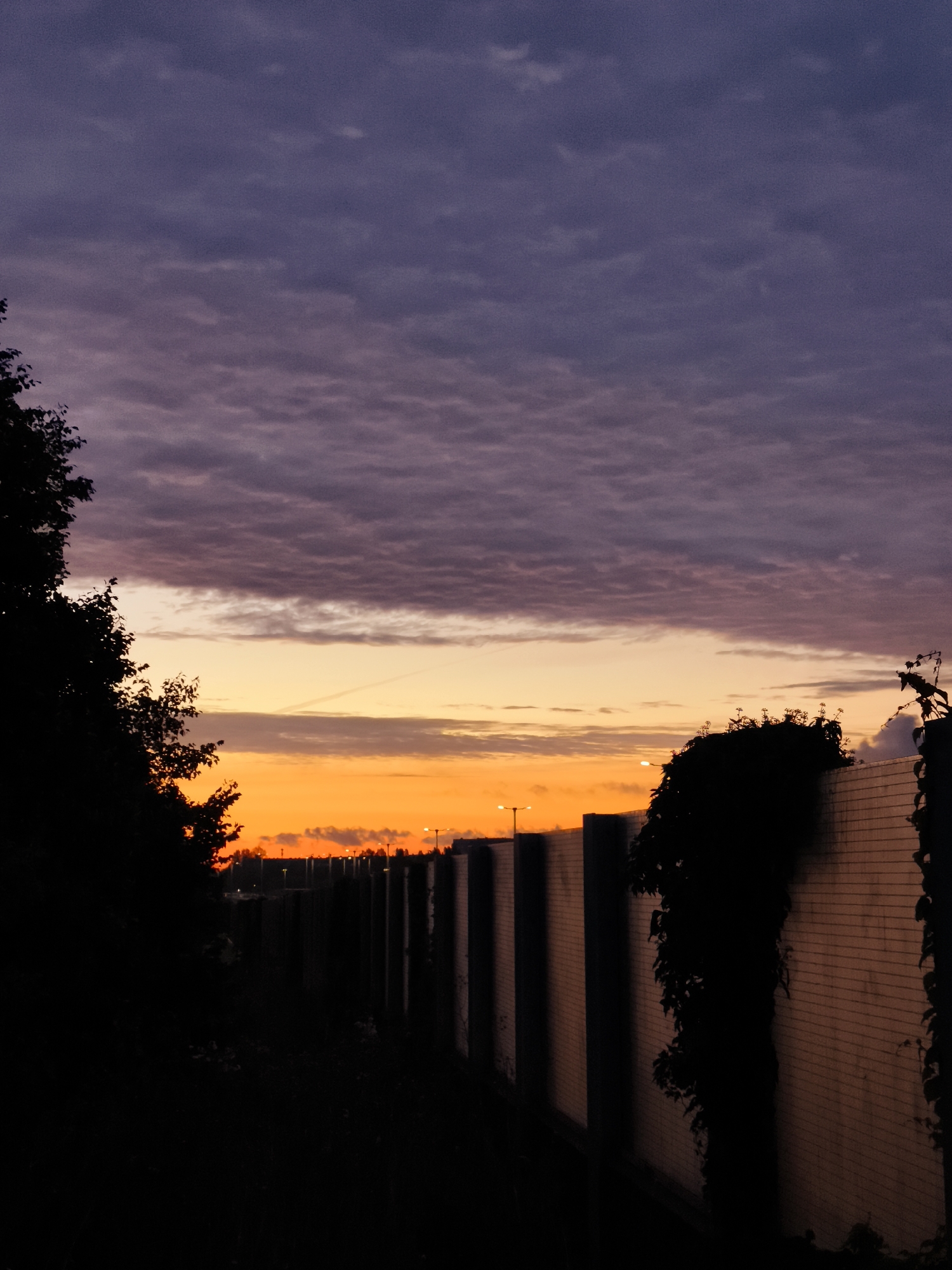 The image depicts a twilight scene with a dramatic sky. The upper portion of the sky is filled with dark purple, textured clouds, transitioning to a bright orange and yellow band near the horizon. A long, straight wall, constructed of light-colored bricks, runs horizontally across the middle ground, creating a strong visual line. The wall is punctuated by regularly spaced, darker rectangular structures, possibly supports or panels. Silhouetted against the vibrant sky are trees on the left and the top of the wall on the right, with some vegetation growing on the wall's surface. Several streetlights are visible in the distance, positioned along the top of the wall, indicating an urban or suburban setting. The overall impression is one of dusk in an outdoor, likely man-made, environment. The location is not identifiable from the visual information present in the image alone.