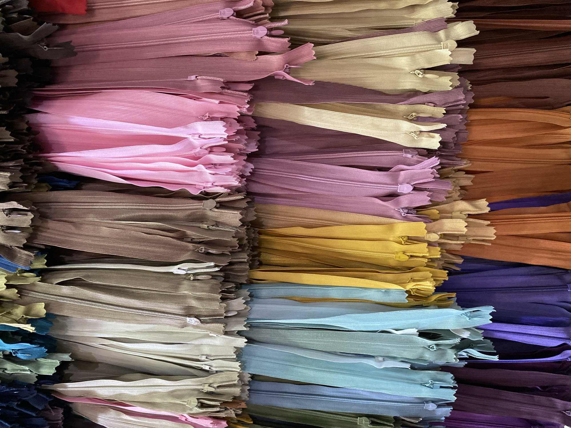 The image depicts a close-up view of a stack of zippers in various colors. The zippers are arranged horizontally and packed tightly together. Colors present include shades of pink, purple, tan, brown, yellow, blue, and green. The environment appears to be indoors. There is no indication of a specific location in Lhokseumawe, Indonesia, or the presence of any people.