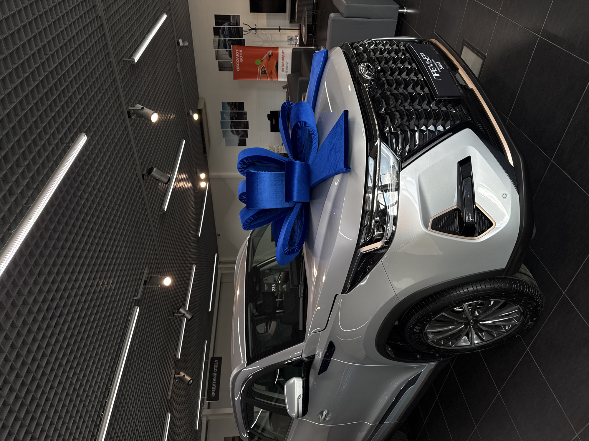 An indoor, eye-level, medium shot shows a light-colored SUV presented as a gift with a large, vibrant blue bow on its hood. The vehicle is the central focus, positioned in a showroom with dark tiled flooring and a grey, modular ceiling equipped with track lighting and fluorescent tubes.
To the left of the vehicle, a sign with the text "КРЕДИТНЫЙ ОТДЕЛ" (Credit Department) is visible, suggesting the setting is a car dealership. In the background, there are posters on the wall, some depicting mountain landscapes and others featuring vehicles. A banner for "АБСОЛЮТ БАНК" (ABSOLUT BANK) is also present. Several pieces of grey seating are arranged in the background, indicating a waiting or lounge area. The overall lighting is bright and even, typical of a commercial showroom. There are no people visible in the frame. The vehicle has a prominent black grille with chrome accents and appears to be brand new. The license plate frame reads "ПРЕМЬЕР SWM".