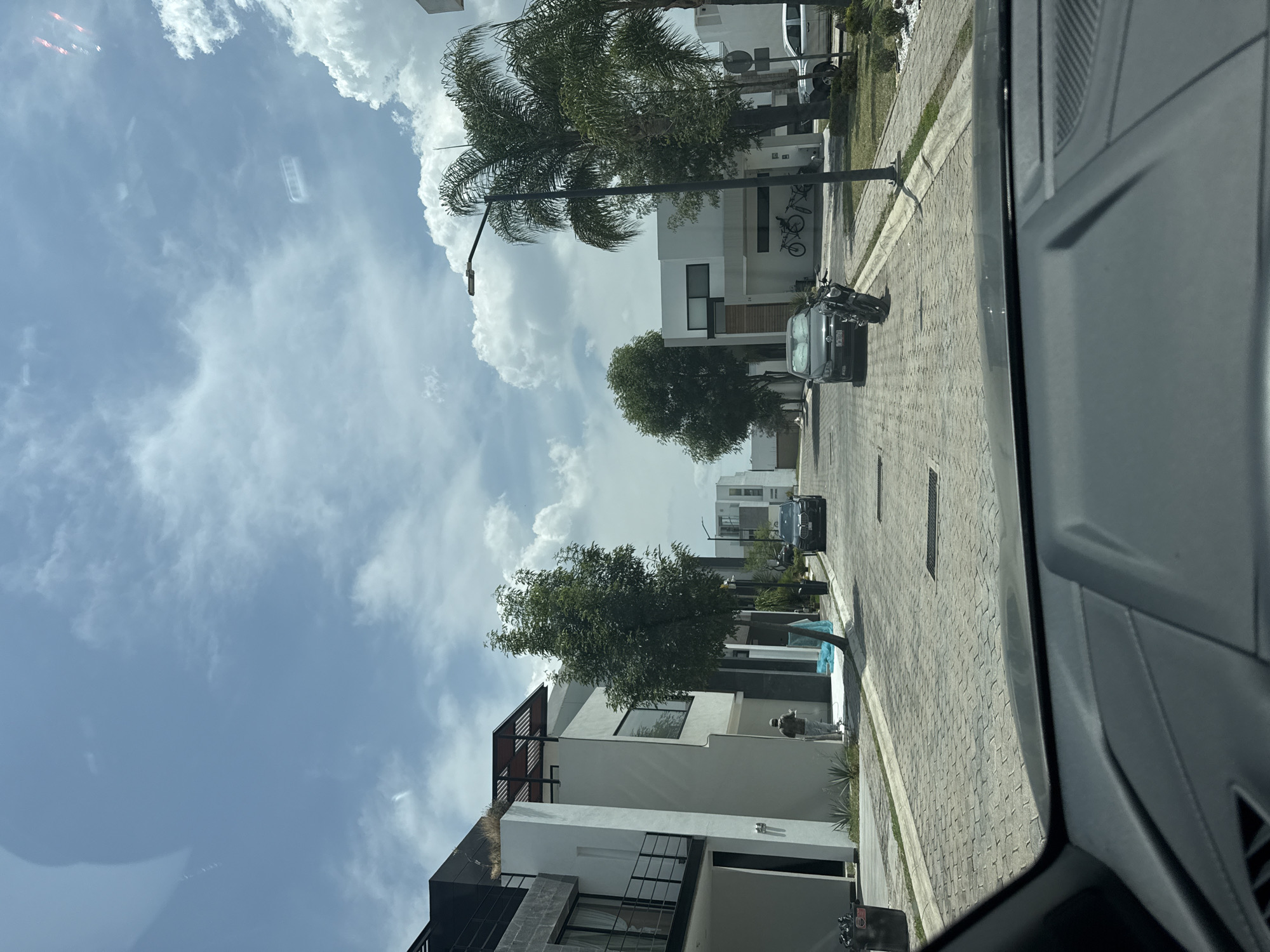 The image, taken from inside a vehicle, displays a residential street in Santa Clara Ocoyucan, Mexico. The street is paved with light-colored interlocking pavers and features modern, two-story houses with contemporary architectural styles, characterized by white and grey facades, large windows, and some balconies or rooftop structures.
The sky is bright and partly cloudy with scattered cumulus clouds. Palm trees and other leafy trees line the street and are present within the properties, suggesting a warm climate. Streetlights are also visible along the road.
Several vehicles are parked on the street, including at least two dark-colored cars and a motorcycle. A person appears to be standing or walking on the sidewalk next to one of the houses. Reflections of the sky and clouds are visible on the vehicle's windshield in the upper left portion of the image. The scene depicts a well-maintained and quiet residential area during daytime. No discernible text is visible.