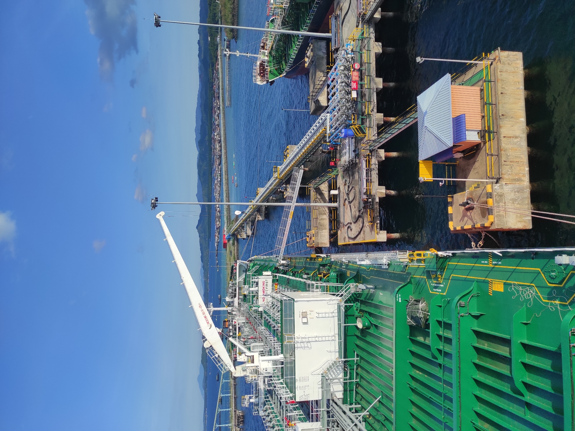 This media file presents a bright, clear-day aerial view of an industrial port in Kunak, Malaysia, depicting a large green tanker ship docked at a concrete pier. The scene is set against a backdrop of deep blue water and a distant coastline featuring mountains and scattered urban development under a partly cloudy sky.
In the foreground, the extensive deck of the green tanker ship is visible, adorned with numerous white structures, intricate piping, and various pieces of machinery. A prominent white crane boom extends from the ship, displaying the text "INGERSOLL SWR 15T@12~25M," indicating its specifications. On the ship's deck, "NO SMOKING" and "SAFETY FIRST" signs are clearly visible, alongside yellow painted lines with text like "FLAP ACCESS POINT KEEP CLEAR," "SAFE AREA," and "DANGEROUS AREA."
The ship is securely moored to a concrete pier, which supports a complex system of yellow, blue, and grey pipelines, access walkways, and stairs. These pipelines connect to the ship, suggesting ongoing or preparatory liquid transfer operations, likely for loading or unloading petroleum products or chemicals. Further down the pier, another, smaller dark-hulled ship is also docked. A small, multi-colored building with a blue roof and orange and blue walls is situated on a section of the pier. Several tall poles with floodlights are scattered throughout the facility, implying capability for continuous operations. The overall impression is of a bustling industrial maritime facility under sunny weather conditions.