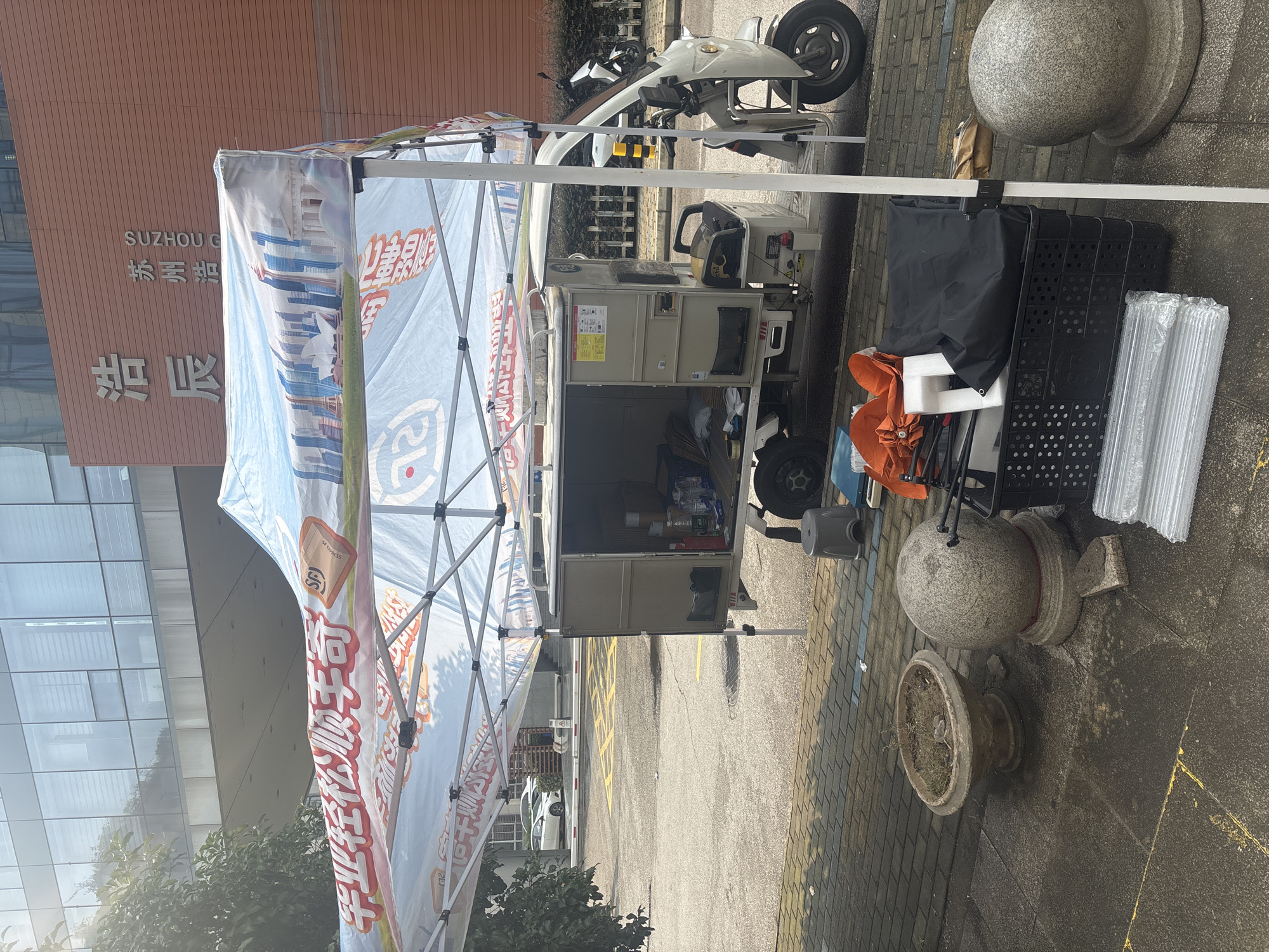 In Suzhou Industrial Park, China, a white electric tricycle is parked beside a collapsible canopy tent. The tricycle has a cargo box at the rear, currently open and containing bottles of water and other items. Various objects are placed on the ground in front of the tent, including a black crate filled with foldable items, a grey stool, and two decorative stone spheres. The canopy tent is adorned with colorful graphics and text, with the building behind it bearing Chinese characters that read "浩辰" and "苏州浩". The environment appears to be an urban outdoor setting with paved ground and a building in the background.