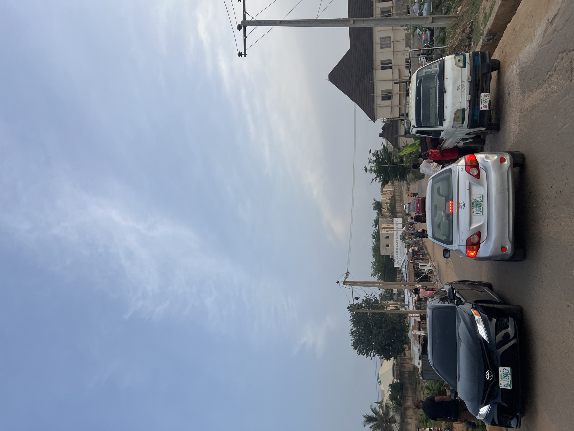 This image depicts a bustling street scene in Abuja, Nigeria, during the daytime under a partly cloudy sky. The scene is dominated by vehicles on a paved road and buildings lining both sides.
In the foreground, a black Toyota Camry sedan, displaying the license plate "NEW NIGERIA KUJ-260ER," is visible. Immediately behind it is a silver Toyota Corolla sedan with the license plate "110 ABUJA ABC-202CL." Further down the road, a white utility van, possibly a Hyundai Porter or similar model, is parked or moving slowly, bearing the license plate "NEW NIGERIA ABC-653XR." Several individuals are seen near the white van, including one wearing a red shirt.
The street is characterized by a mix of buildings, some appearing residential and others commercial. One building in the mid-ground shows scaffolding, indicating construction or renovation. Utility poles with numerous overhead wires are prominent features along the roadside. Various people are visible walking on the street's shoulders, suggesting pedestrian activity or informal market operations. Trees and other green foliage are scattered throughout the urban landscape. A sign with the text "URBAN SHE" is visible on a building in the background. The overall impression is of a lively urban or semi-urban environment with ongoing daily activities and traffic.