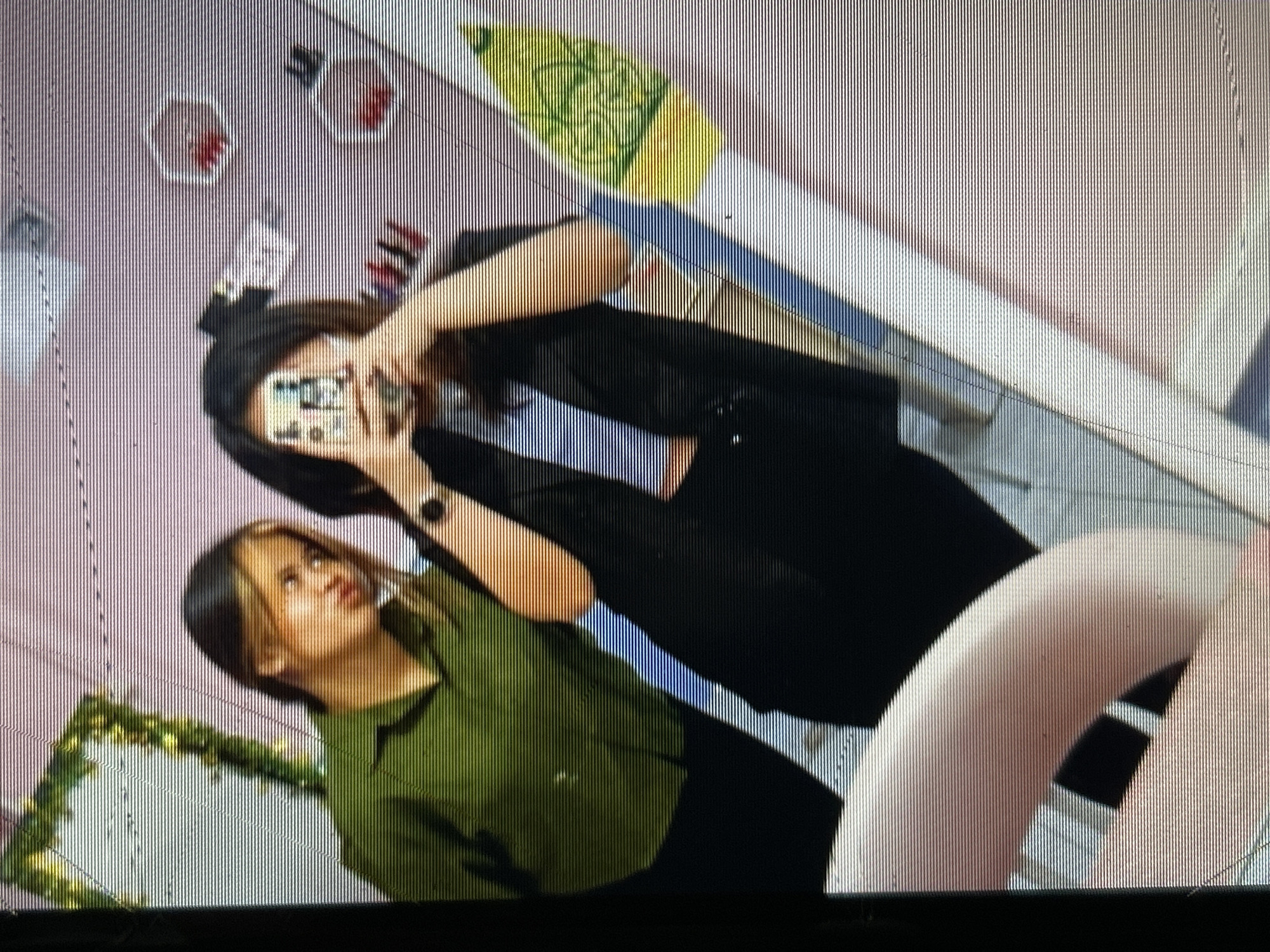 The image shows a reflection in a mirror of two young women. The woman on the left is wearing a green collared shirt and has her hair pulled back. She is looking towards the right with her lips pursed. The woman on the right is wearing a black blazer over a light blue top. She is holding a smartphone in front of her face, seemingly taking a selfie. Her hair is dark and pulled back. The background appears to be a room with a pink wall. There are some decorative items on the wall, including what looks like small hexagonal shelves with red objects inside. A green garland with leaves is visible in the upper left corner of the reflection. The image is characterized by a strong horizontal line pattern, indicative of a low-resolution capture or display artifact. The context suggests this photo was taken in Depok, Indonesia, likely in a personal setting like a bedroom or dorm room.