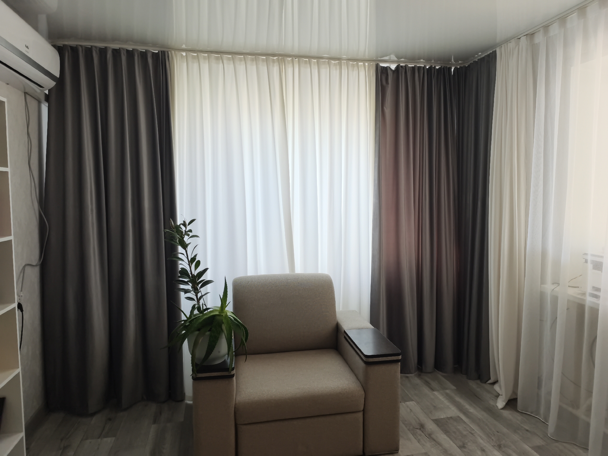 This is a medium shot, eye-level, indoor photograph of a sparsely furnished room. The room features a light-colored sofa, dark gray curtains on the left and right, and sheer white curtains in the center. A potted plant sits on a small pedestal to the left of the sofa. An air conditioning unit is mounted high on the wall above the curtains. The floor is made of light-colored wood-effect laminate. The overall impression is one of a living space that is functional but not yet fully decorated. The city of Novo-Oktyabrsky, Russia is mentioned as the location, but there are no specific visual cues to confirm this.