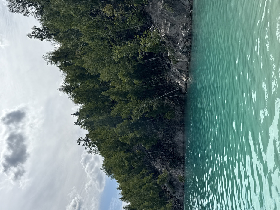 The image displays a panoramic view of the Druzhba Quarry lake located near Druzhba, Sumy Oblast, Ukraine. The foreground is dominated by a body of water exhibiting a distinct turquoise hue, with numerous surface ripples visible. The water borders a steep, rocky embankment on the right side of the frame, characterized by dark grey and black rock formations and scattered scree. Above the embankment, a dense forest of deciduous trees, predominantly green-leafed species, covers the sharply rising hillside. The trees extend vertically, some appearing angled on the slope. The sky above is partially cloudy, featuring grey and white clouds interspersed with brighter patches. No individuals, animals, or man-made structures are present within the scene.