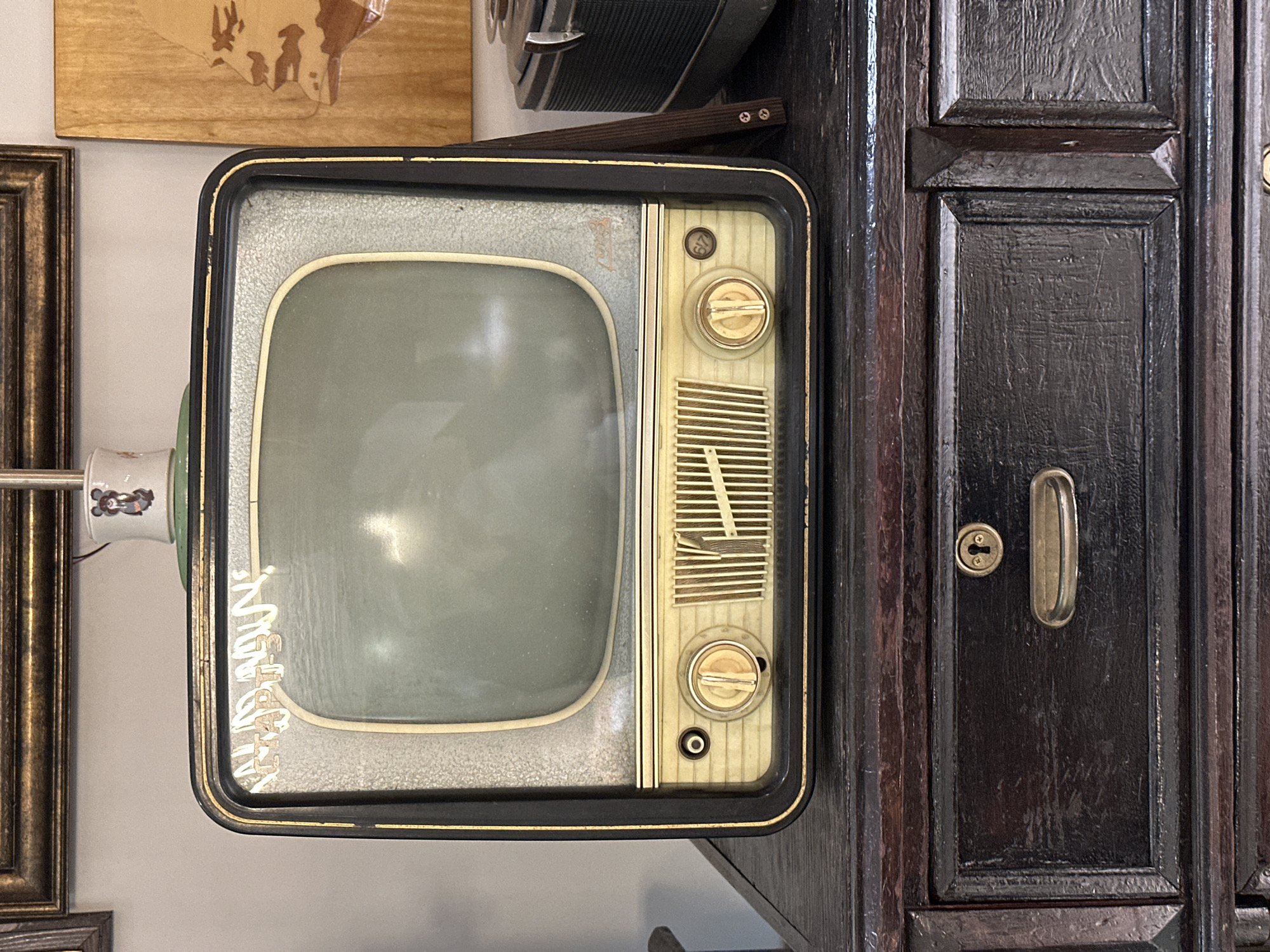 The image shows a vintage television set placed on top of a dark wooden dresser. The television is black and gold with a cream-colored bezel around the screen. The screen itself is a muted green. There are two round knobs and a grille on the front of the television. To the left of the television, there is a framed picture on the wall, and to the right, a wooden carving of a face. The dresser has two drawers, one with a brass handle and keyhole. The scene appears to be indoors, likely in a home or a shop. The city of Blagoveshchensk, Russia is not visible in the image, nor are there any details that would place it specifically in that location. The overall impression is one of retro decor.
