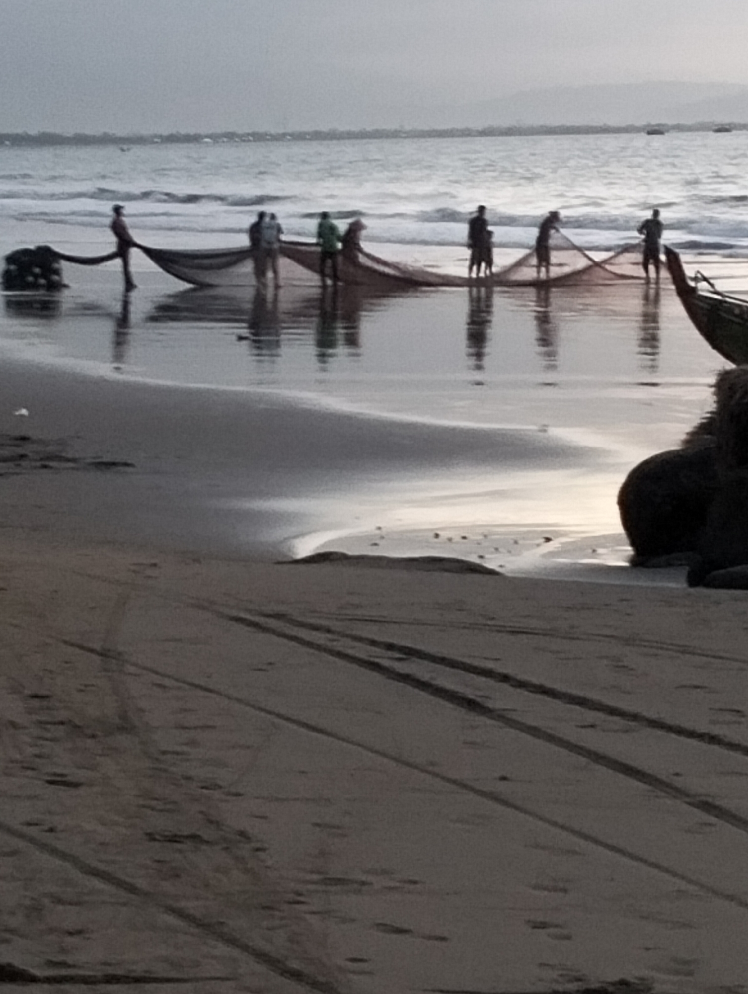 In Peuneulet Baroh, Indonesia, a group of fishermen are working on a beach with a large fishing net spread across the shallow water and sand. The sky is overcast, and the water appears choppy with waves breaking on the shore. The sand in the foreground has tire tracks imprinted on it. In the background, the ocean stretches out to the horizon, with a distant coastline visible. A dark object, possibly a boat, is partially visible on the right side of the frame.