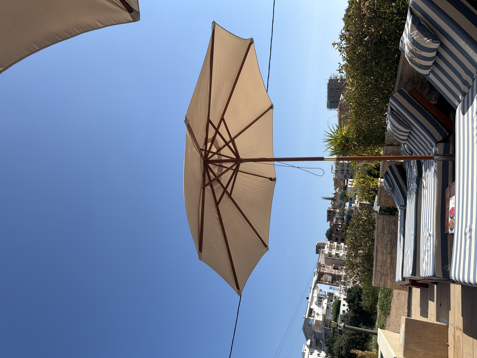 The image depicts a bright, sunny day with a clear blue sky dominating the upper portion. In the foreground, several lounge chairs with blue and white striped cushions are arranged on a paved area, suggesting a poolside or patio setting. A large, light-colored umbrella with wooden ribs is open and centered in the frame, providing shade. Behind the lounge chairs, a low stone wall topped with dense green foliage separates the seating area from a backdrop of buildings. The buildings are a mix of architectural styles, with several appearing to be apartment complexes or hotels, situated on a hillside. Some buildings are white, while others feature colorful facades. Construction scaffolding is visible on one of the buildings in the distance, indicating ongoing development. A power line runs horizontally across the mid-section of the image, partially obscured by the umbrella. The scene is consistent with a resort or residential area in a coastal or hilly town. The location is identified as Pečurice, Montenegro.