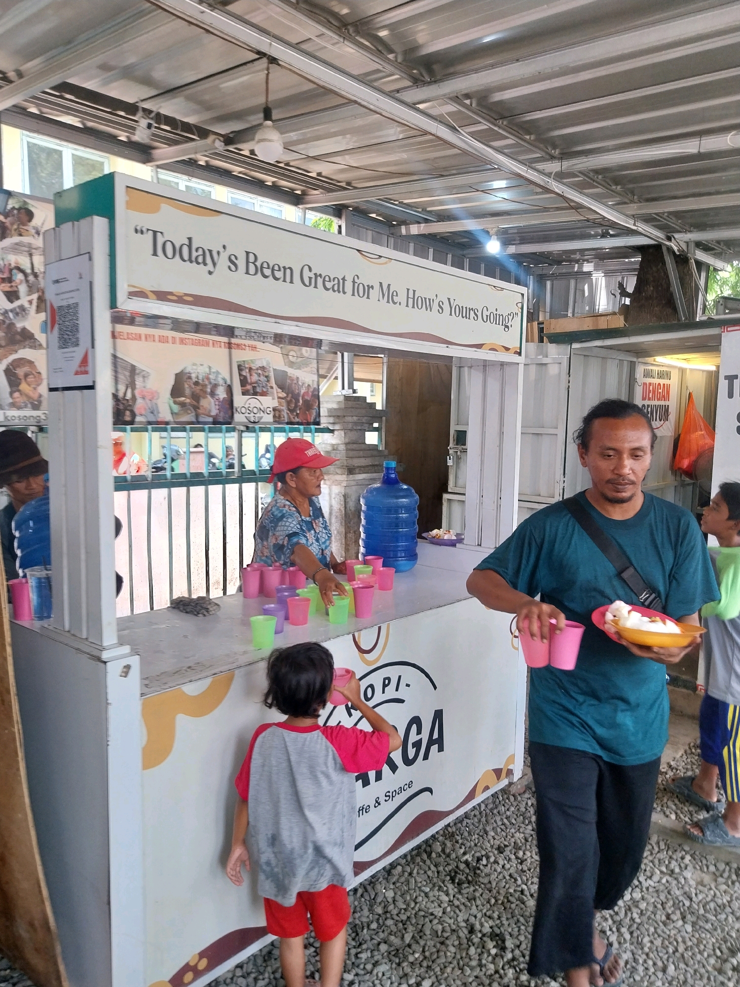 An exterior shot captures a food stall under a corrugated metal roof with exposed beams and lights. A large sign on the stall reads "Today's Been Great for Me. How's Yours Going?". In the foreground, a man in a teal t-shirt and black pants holds a plate of food and two pink cups. To his right, a young boy in a green shirt and striped shorts walks away from the stall. Behind the counter, a woman wearing a red cap and a patterned shirt serves customers, with stacks of pink and green plastic cups on the counter. A young child in a gray and red shirt stands at the counter, facing away from the camera. A large blue water jug is visible behind the counter. The ground is covered in gravel. The stall appears to be set up in a public area, possibly a market or outdoor dining space. The signage and context suggest it's a food and beverage service.