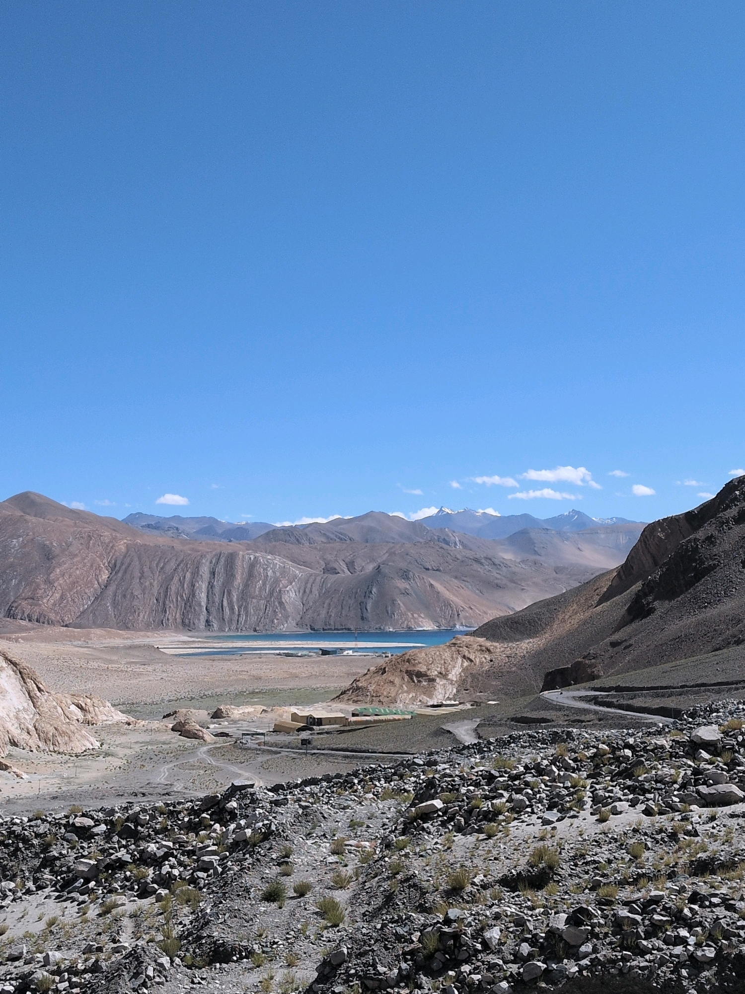 An expansive, high-angle, eye-level shot captures a desolate, mountainous landscape under a clear blue sky in an unknown location in India. The foreground is dominated by a rough, rocky terrain with sparse, dry vegetation. A winding, grey road curves upwards along the right side of the frame, leading into the distance. In the midground, a vast, arid valley stretches out, dotted with a few small, light-colored buildings and tents, some with green roofs. A body of water, appearing as a calm, blue lake or large pond, is visible in the distance, nestled between the valley floor and the surrounding mountains. The background is defined by imposing, arid mountains with textured, eroded slopes, rising towards the horizon where more distant, snow-capped peaks are faintly visible. The overall impression is one of stark, natural beauty and isolation.