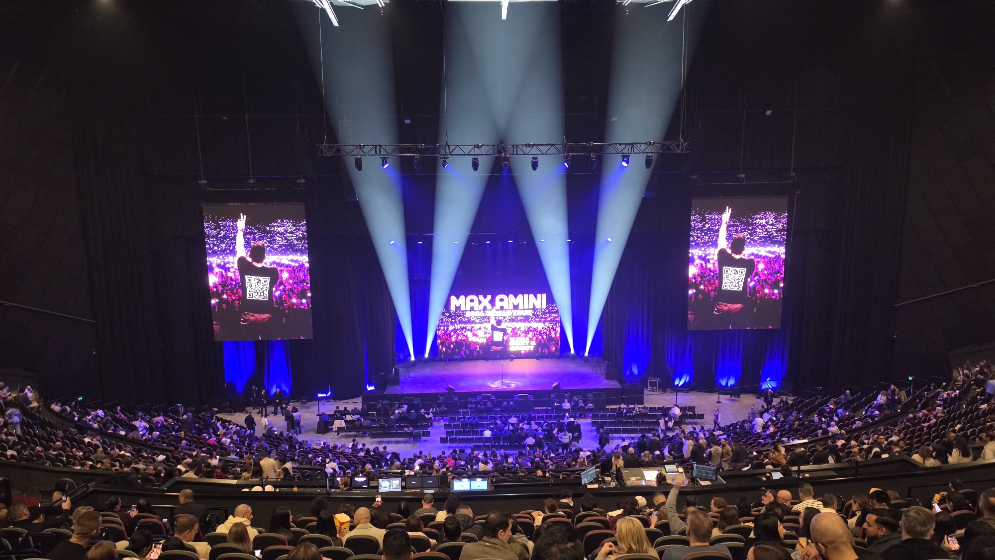 The image depicts a large indoor performance venue, likely a theater or arena, filled with a seated audience. The stage is set with a backdrop displaying the name "MAX AMINI" and an image of a performer with arms raised, superimposed on a crowd. Spotlights from above illuminate the stage. Two large screens flank the stage, each showing the performer's image. The audience is seated in tiered rows facing the stage, with some individuals visible in the foreground. The overall atmosphere suggests a live entertainment event. The specific location in Sydney, Australia, cannot be determined from the image alone.