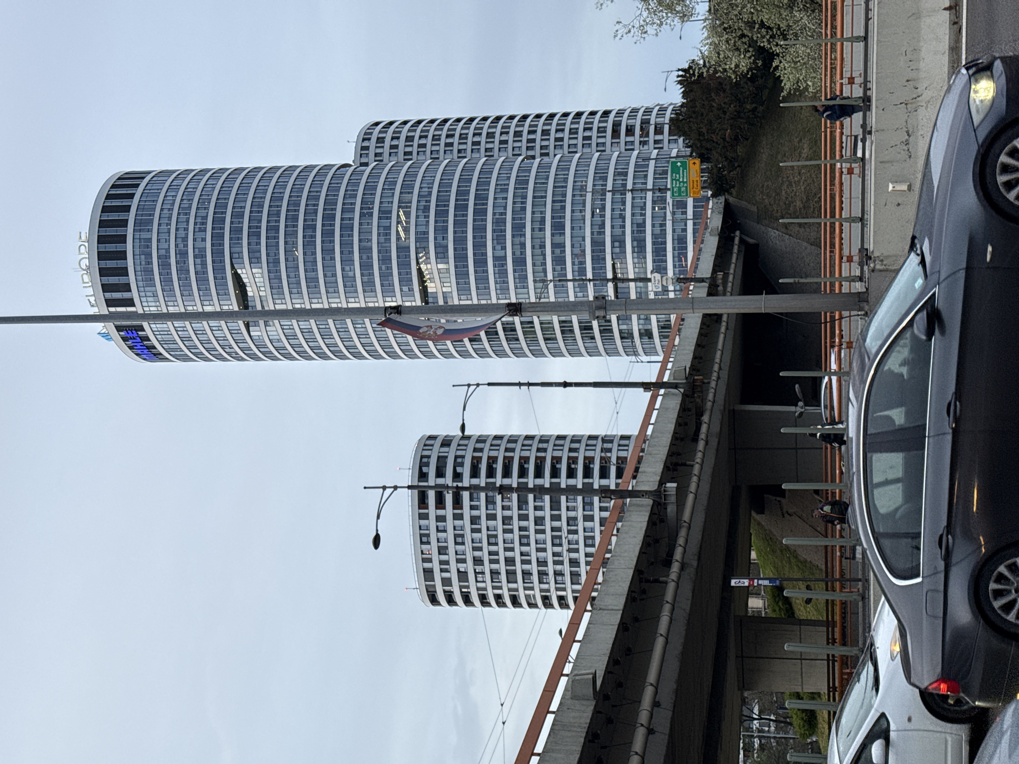 The image shows a scene in Belgrade, Serbia, featuring the Genex Tower complex. Two cylindrical towers with horizontal white stripes are visible. A Serbian flag is hanging. A bridge or overpass spans above a road, with cars, including a black sedan and a white car, visible on the right side. A street lamp is in the foreground. Vegetation is seen growing near the bridge. There appear to be people near the bridge walkway. The sky is overcast.