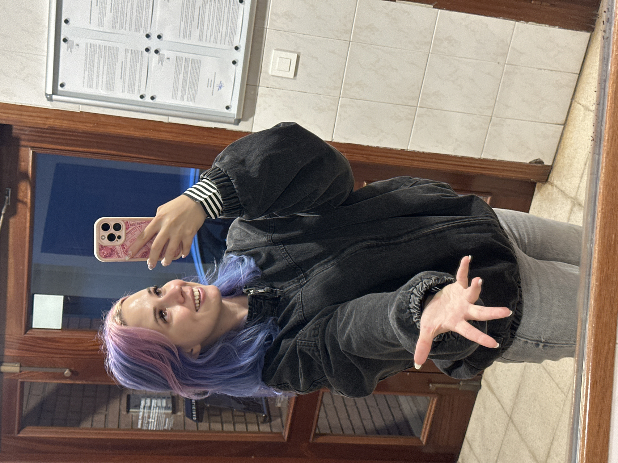 The image depicts a young woman taking a mirror selfie in Gijón/Xixón, Spain. She has long, wavy hair dyed in vibrant shades of purple and pink, with lighter roots. Her face is turned slightly to the right (viewer's left), displaying a wide, joyful smile with her eyes cast upwards. She is wearing an oversized dark gray/black denim bomber jacket with a zippered front, ribbed cuffs, and collar. The cuff of a black and white striped long-sleeved shirt is visible on her left wrist. She is also wearing light gray jeans. Her right hand holds a light pink, patterned smartphone, positioned to capture the reflection. Her left hand is extended forward towards the viewer, palm slightly open with fingers splayed. Her fingernails are manicured with a light/white polish.
The mirrored background reveals a tiled wall on the right side, composed of light beige rectangular tiles with dark grout lines. Mounted on this wall is a silver-framed notice board containing multiple sheets of paper secured by visible pins. Below the notice board, a single square white light switch is present. To the left, a dark wooden door frame with a glass panel reflects a darker, indistinct background, possibly featuring brickwork and a partially legible sign. The floor visible in the reflection is covered with light-toned square tiles. The lower wooden frame of the mirror itself shows small dark specks of dirt or debris.