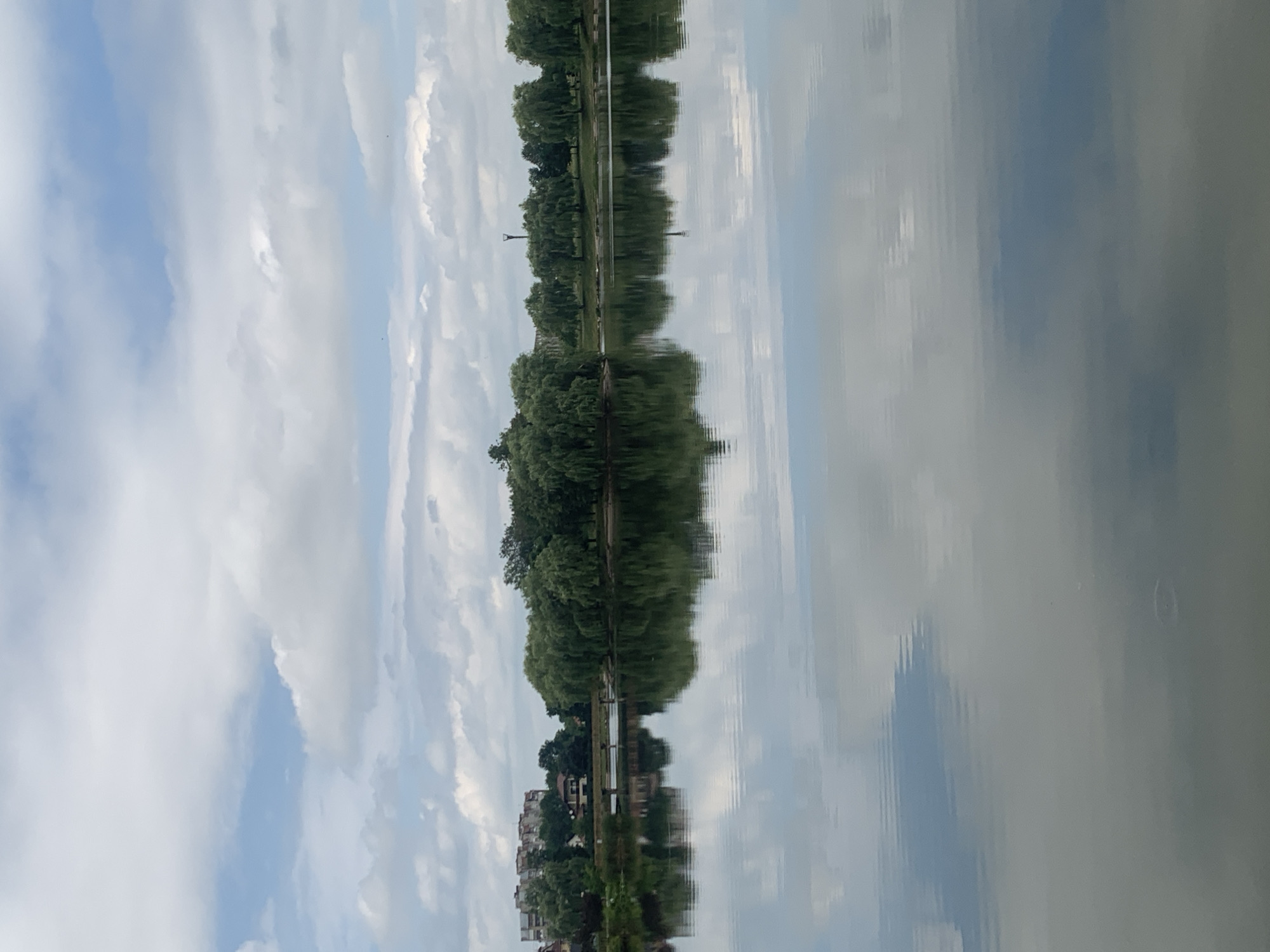 A serene body of water reflects a cloudy sky and a lush green treeline with buildings in the distance. The water's surface is calm, with subtle ripples creating a distorted mirror image of the environment above. The treeline is dense, featuring a variety of trees, including willows with their characteristic drooping branches. Behind the trees, several multi-story buildings are visible, suggesting an urban or suburban setting. The sky is partially overcast, with large, fluffy white clouds interspersed with patches of blue. The overall impression is one of peacefulness and natural beauty. This image was captured in the city of Kolomyia, Ukraine.
