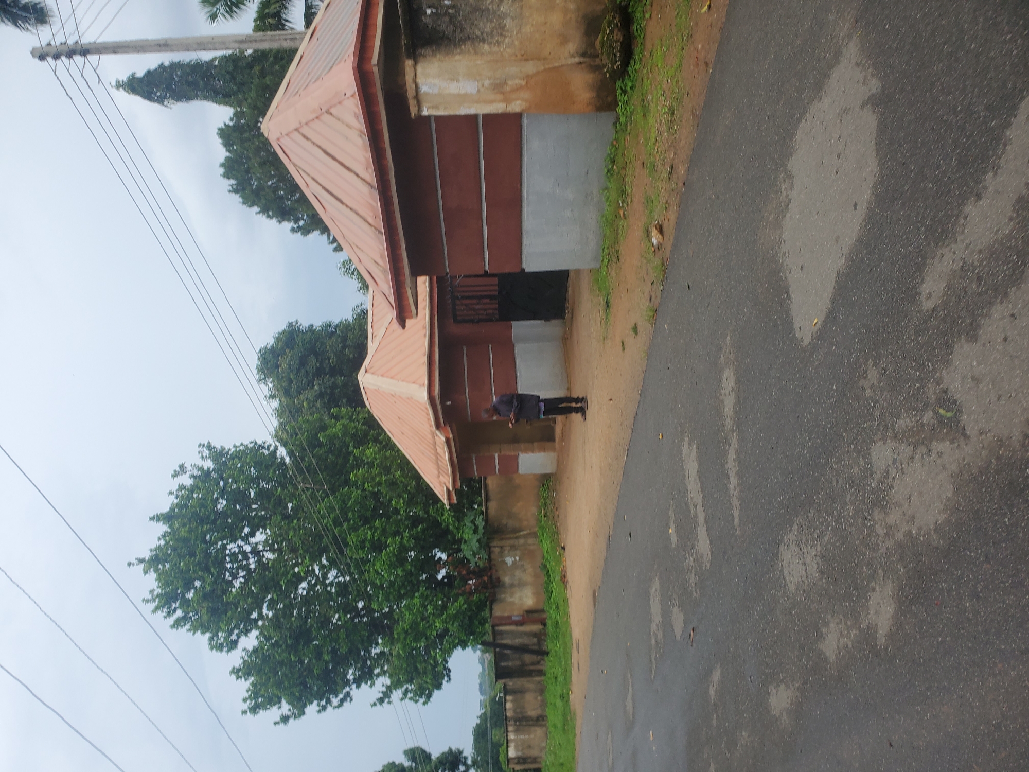 The image depicts a street scene in Jos, Nigeria. A person stands near a building with a reddish-brown exterior and a corrugated metal roof. The building appears to be part of a compound or an entranceway, with a black metal gate visible behind the person. The street in the foreground is paved with asphalt and shows signs of wear, with several wet patches indicating recent rain. Beyond the building, a low, concrete wall with some vegetation growing along its top stretches into the background. Large trees with lush green foliage are scattered throughout the scene, and utility poles with wires are visible against an overcast sky. The overall impression is one of an ordinary urban or semi-urban environment.