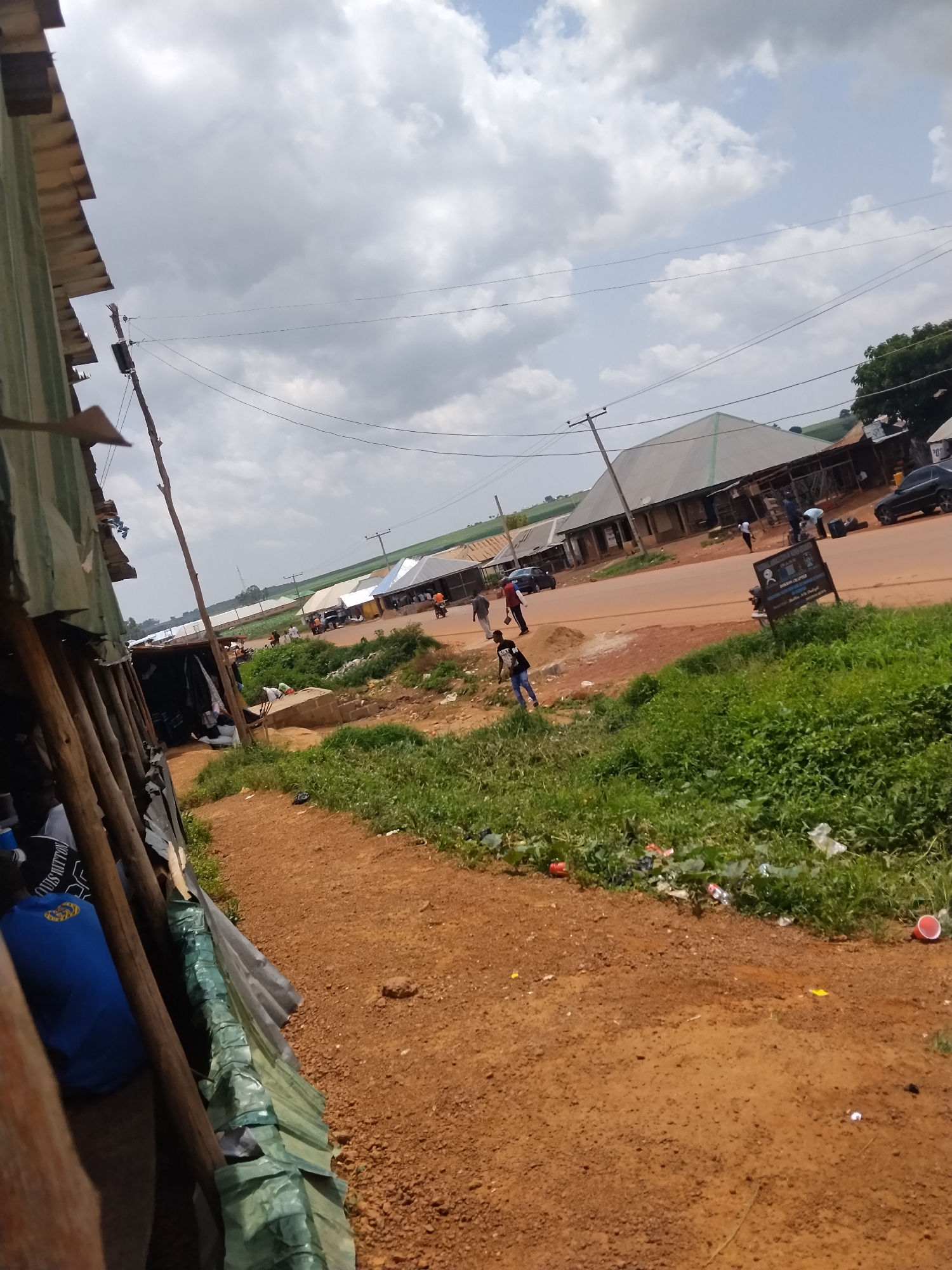 The image depicts a rural scene in Durumi, Nigeria, on a cloudy day. The foreground is dominated by a dirt path and an area of lush green grass bordering a road. Various litter items, including plastic bottles and red cups, are scattered in the grass. On the left side of the frame, a makeshift structure constructed from corrugated metal and wooden poles is visible, with what appears to be fabric or plastic sheeting draped over it. Inside this structure, a blue bag with a yellow logo is partially visible.
Across the road, several low-rise buildings with corrugated metal roofs are present. Some of these buildings appear to be shops or stalls. A few people are seen near these buildings and on the road. One individual in the mid-ground is wearing a white shirt and dark pants, and another is in a white shirt and dark pants. Further down the road, a few cars and motorcycles are parked or moving. A utility pole with power lines stands prominently in the middle of the scene. In the background, green fields extend towards a treeline, and more buildings are visible, suggesting a small settlement or village. The overall impression is one of a community along a rural road, with elements of commerce and everyday life.