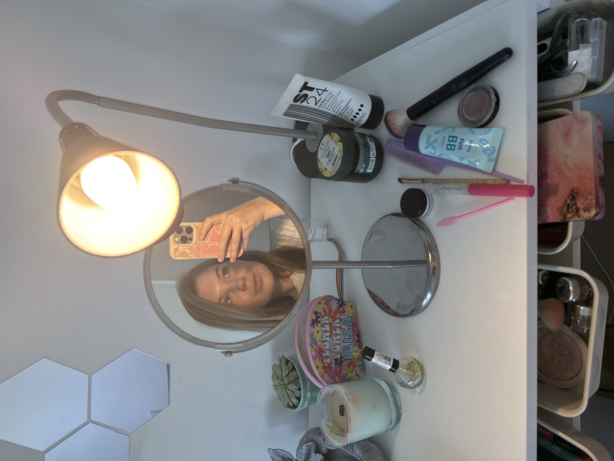The image presents an overhead shot of a vanity area or desktop. A gooseneck lamp illuminates the scene, its light reflecting off a round vanity mirror. A person is visible in the mirror, holding a phone up to take a selfie. The countertop is cluttered with various cosmetic and personal care products, including a tube of hair gel with the text "ST 24 EXTRA ГЕЛЬ для волос" in Russian and "ESTEL", a jar of moisturizer, a BB cream tube labeled "Petit", makeup brushes, a small comb, and other cosmetic items. There's also a small succulent, a decorative bag that says "Flower Power Ibiza," a candle, and a nail polish bottle. The right side of the frame features a section of vertical white drawers or shelves, likely part of a vanity organizer, filled with additional cosmetic items. The background features hexagonal mirror tiles. The overall scene suggests a personal grooming or makeup application setting. The location is indicated to be Vozdovac Urban Municipality, Serbia.