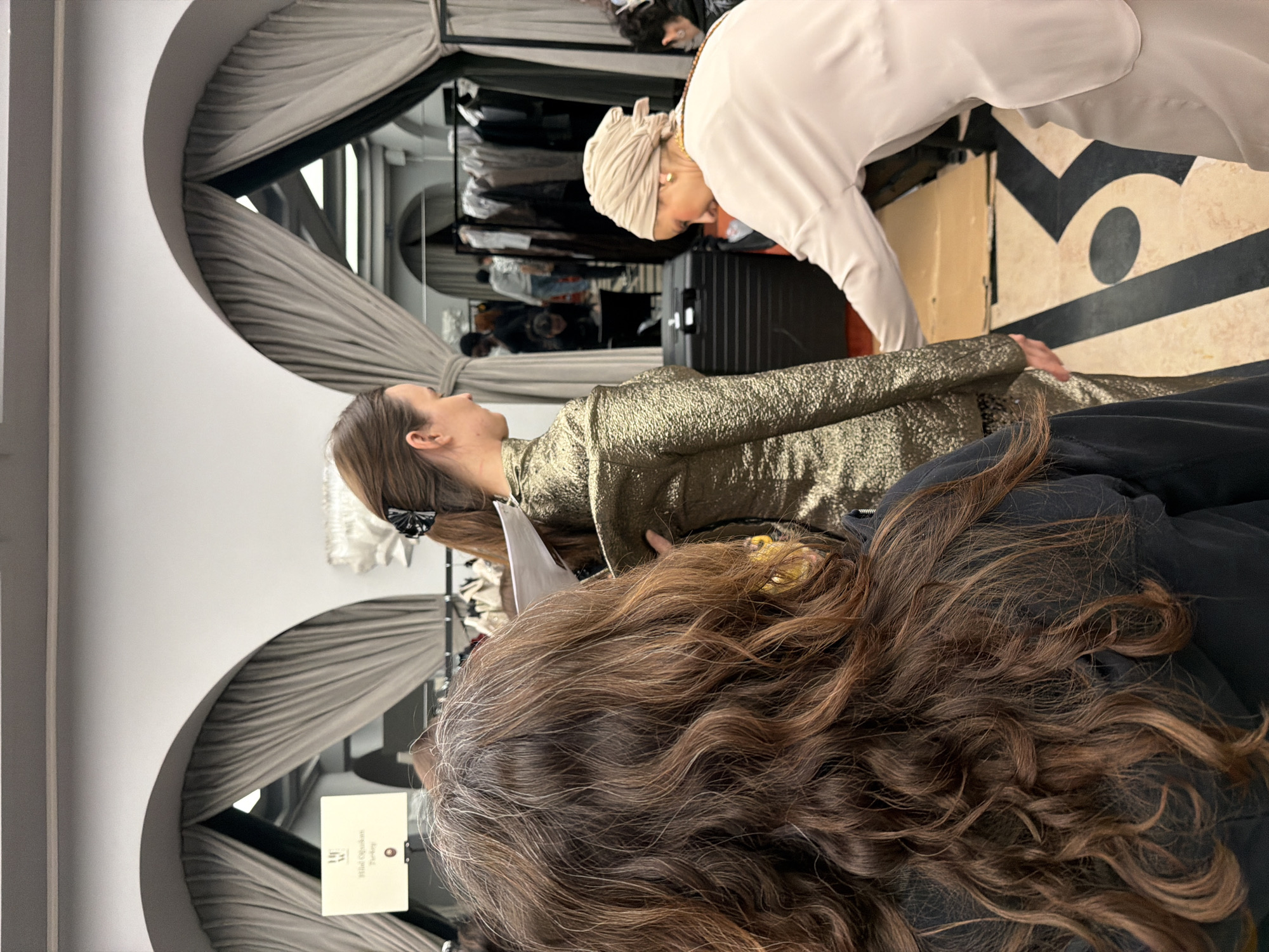 A vertically oriented image captures a behind-the-scenes moment, possibly in the fashion industry, likely in Paris, France, based on the presence of people and objects. The scene is indoors, possibly a fitting room or backstage area, as indicated by the presence of wardrobes and clothing racks with garments.
Several people are present. A woman with dark brown hair is in the foreground, facing away from the camera, with the back of her head and shoulders taking up a significant portion of the bottom of the frame. Partially visible is another woman dressed in what appears to be a shimmering gold garment. A third woman, wearing a beige head covering and a light-colored outfit, is leaning over her. The scene suggests that the gold-clad woman is being fitted or adjusted in her clothing.
The setting includes arched openings covered with gray curtains, and mirrors that show a larger dressing room-like area with clothing racks, suitcases, and the reflections of additional people. There is a sign hanging from one of the arches. The floor has a geometric pattern.