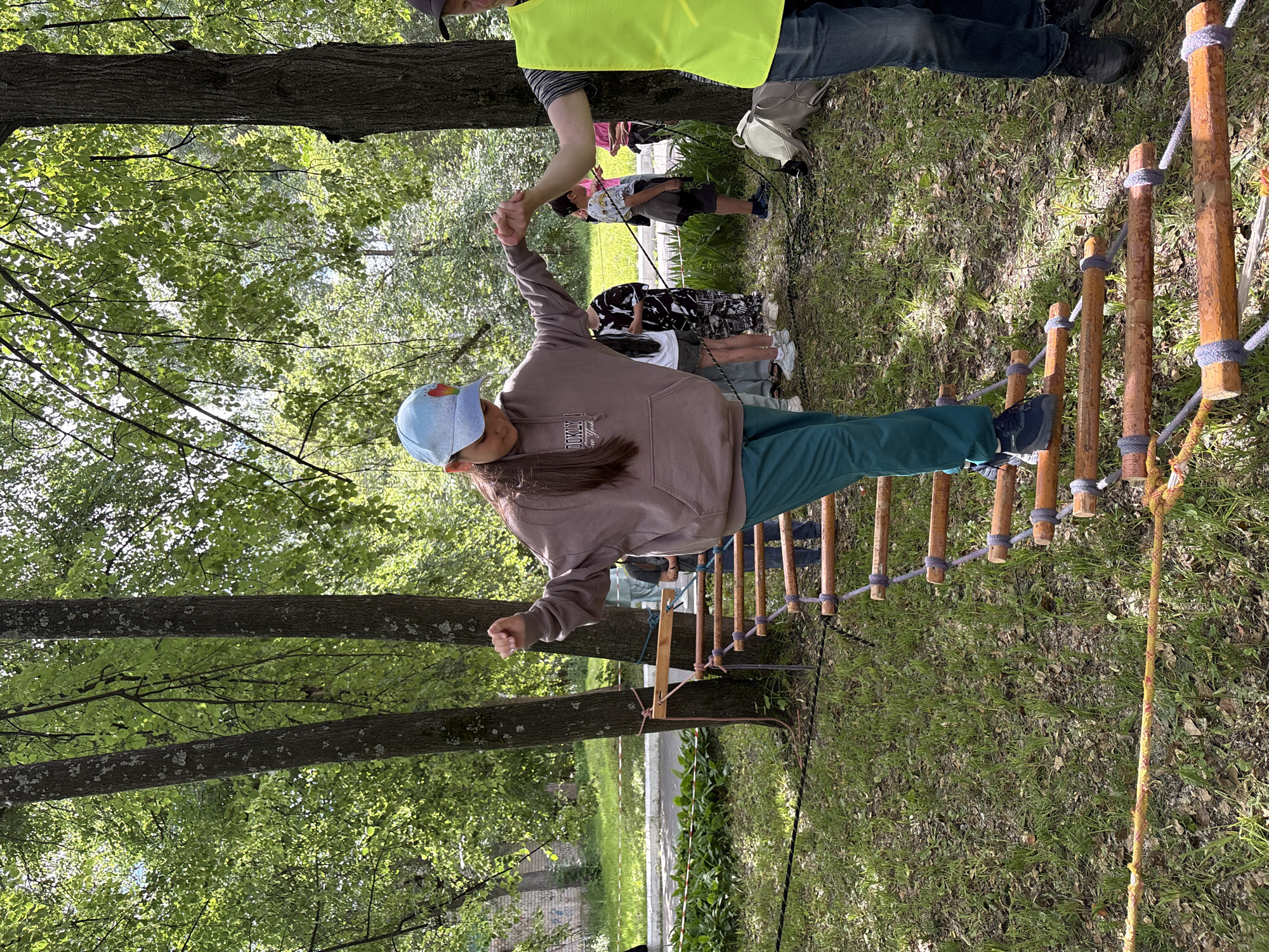 The image depicts an outdoor rope bridge obstacle course, likely in a park or wooded area within the city of Chaikovsky, Russia. A young person wearing a blue cap, a brown hoodie, and green pants is actively crossing the rope bridge, holding hands with another person who is partially visible on the right side of the frame, wearing a bright yellow vest. Several other individuals, appearing to be children and adults, are standing in the background, observing or waiting. The environment is characterized by trees with green foliage, grass, and sunlight filtering through the leaves. The rope bridge itself is constructed from wooden planks and thick ropes.